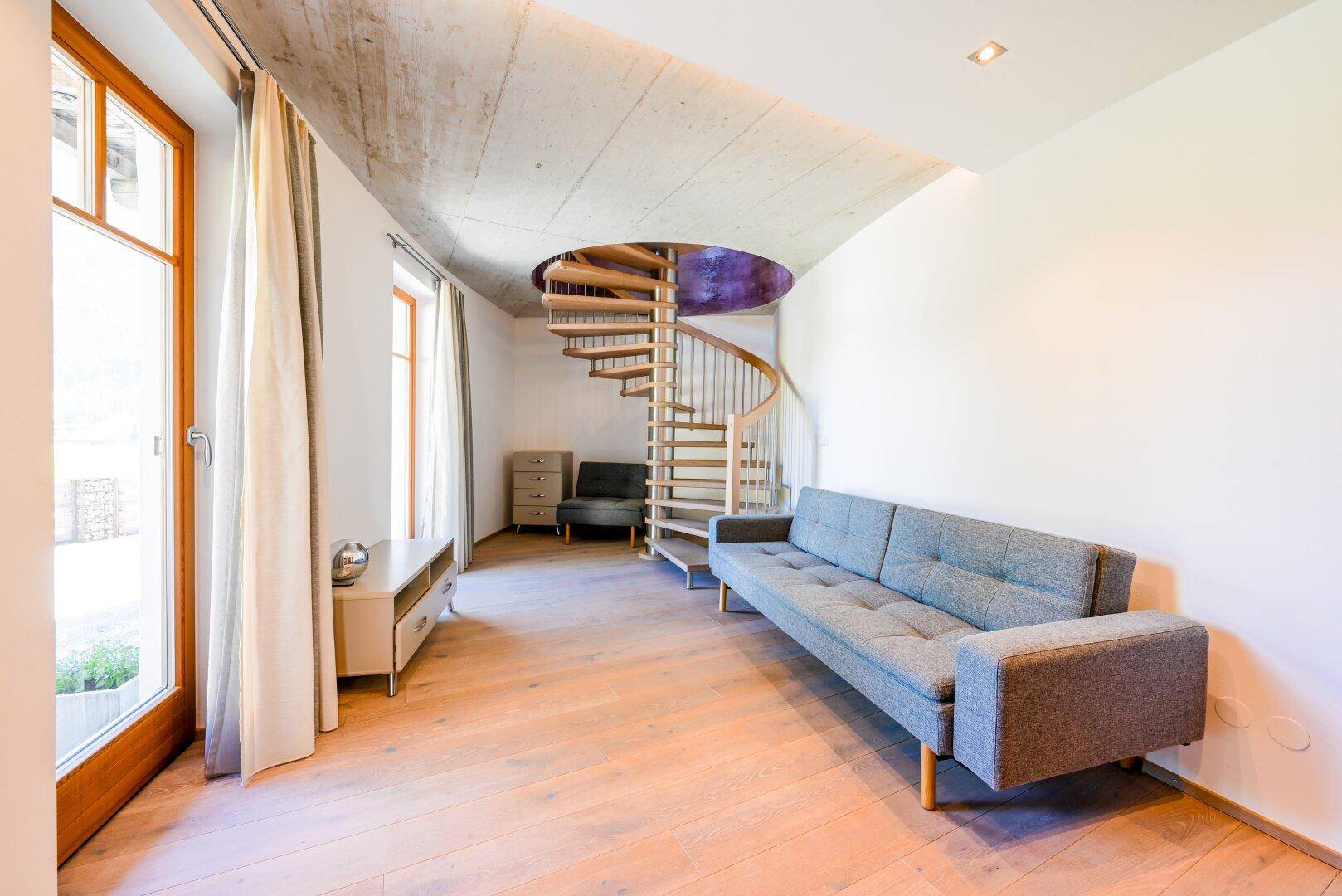Heller Wohnbereich mit Wendeltreppe, Sofa und großen Fenstern, die viel Licht spenden.