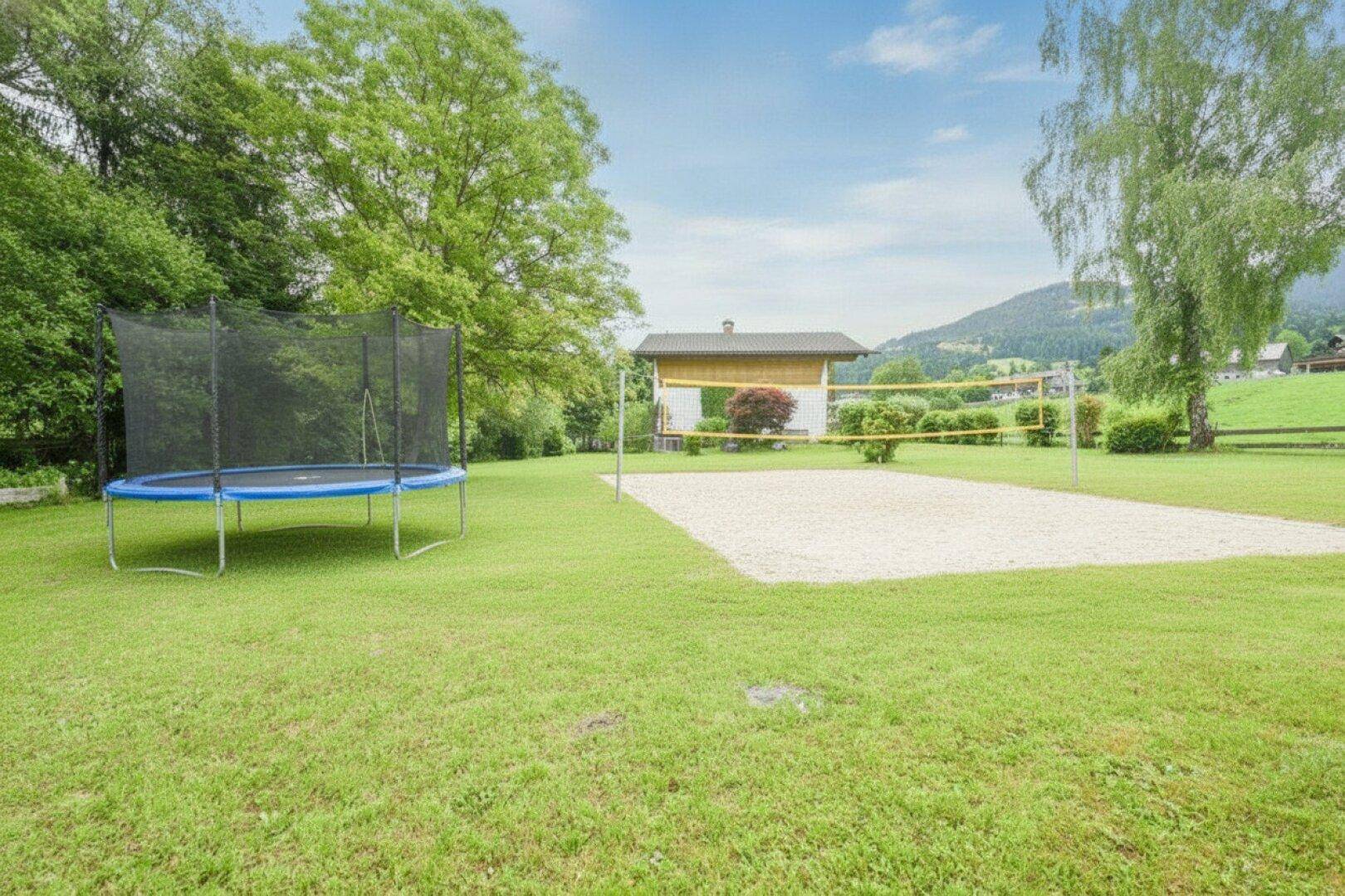 Großer Gartenbereich mit Trampolin und Beachvolleyballfeld, umgeben von viel Grün.