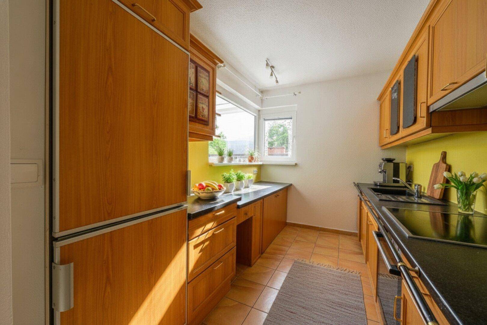 Funktionale Küche mit Holzschränken, dunklen Arbeitsplatten, Fliesenboden und Fenster.