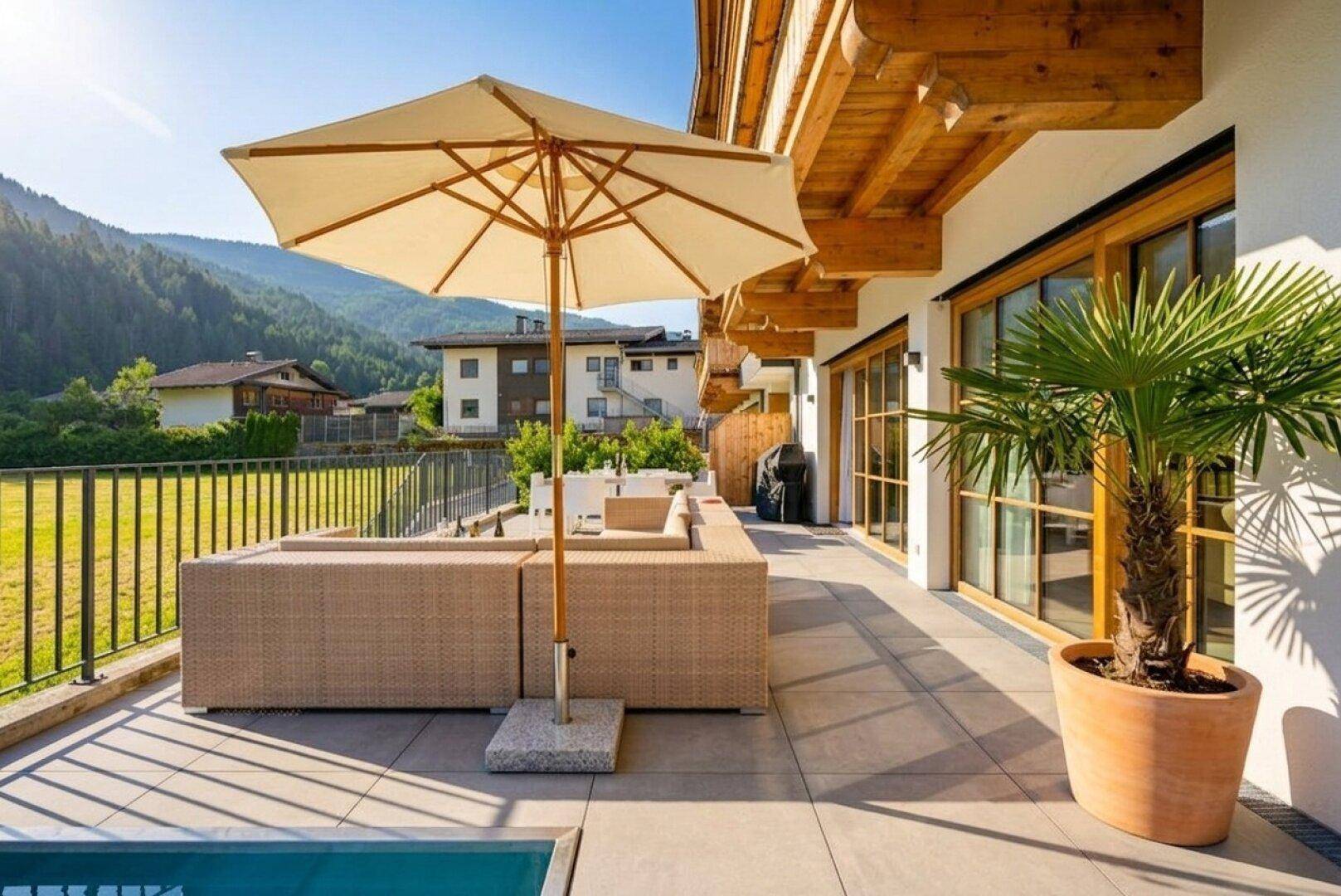Sonnige Terrasse mit bequemen Loungemöbeln, Sonnenschirm und Blick auf die umliegende Berglandschaft.