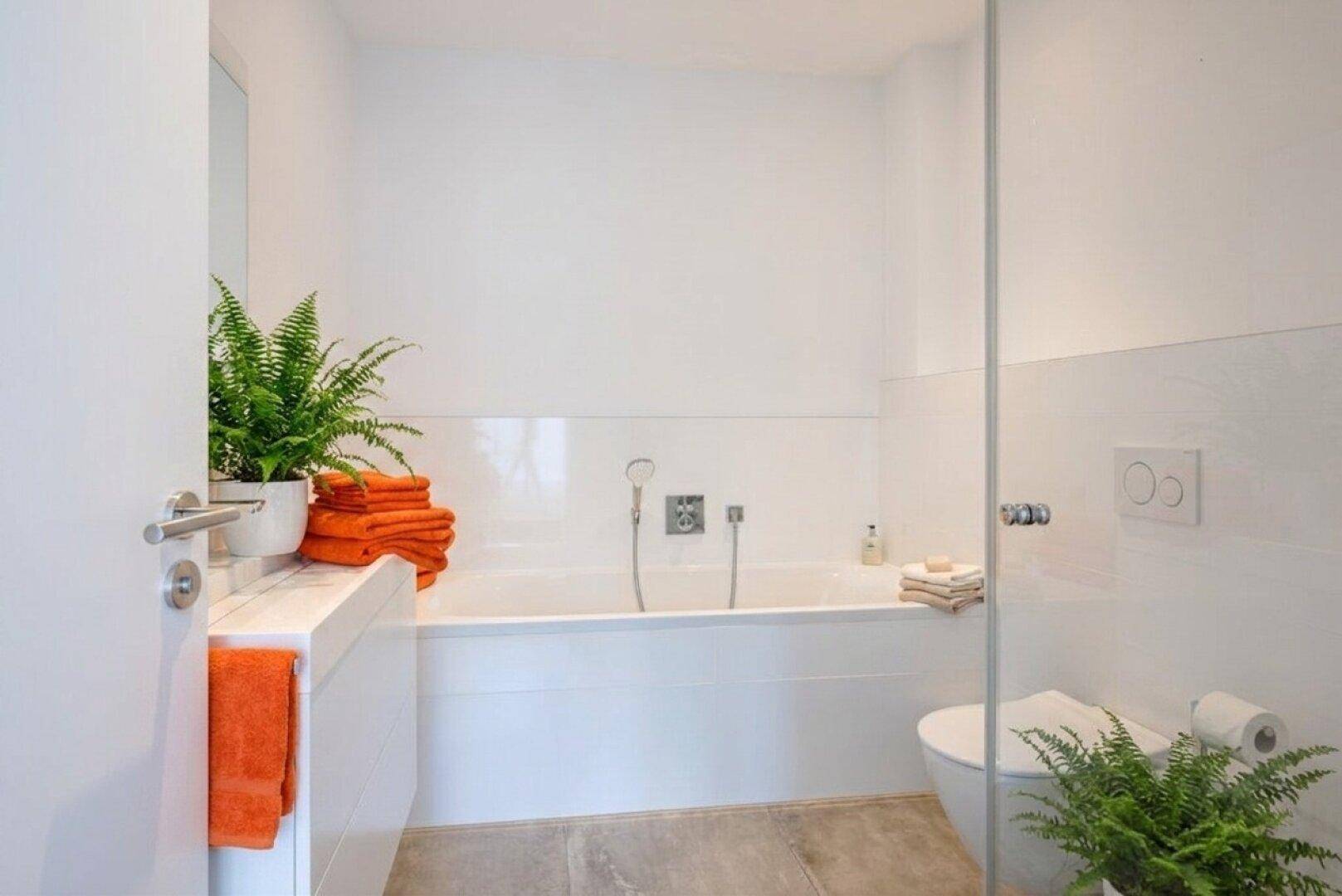 Modernes Badezimmer in Weiß mit Badewanne, Wand-WC und frischen Pflanzenakzenten.