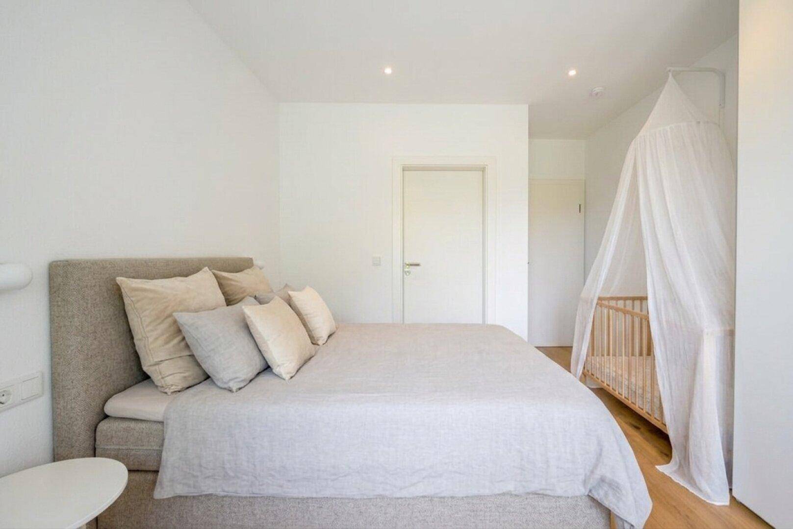 Ruhiges Schlafzimmer mit Doppelbett und Babybett, in hellen, neutralen Farbtönen gehalten.