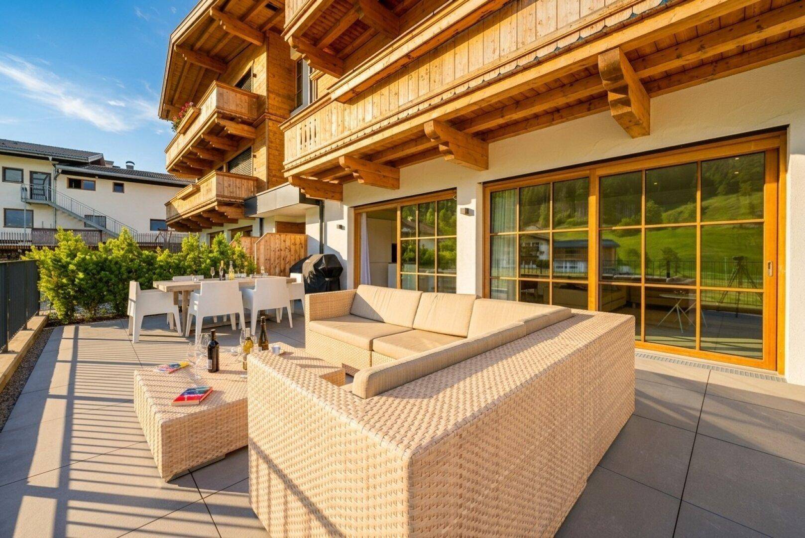 Große Terrasse mit Rattan-Lounge und Esstisch, umgeben von grüner Natur und Bergen.