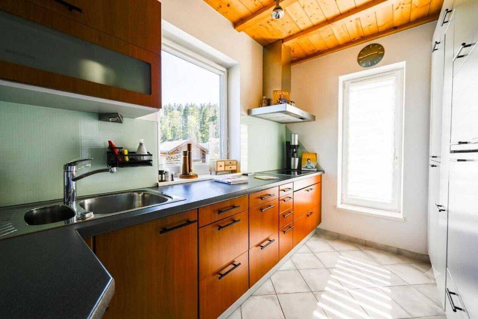 Funktionale Küche mit Holzschränken, Fliesenboden und Fenster mit Waldblick.