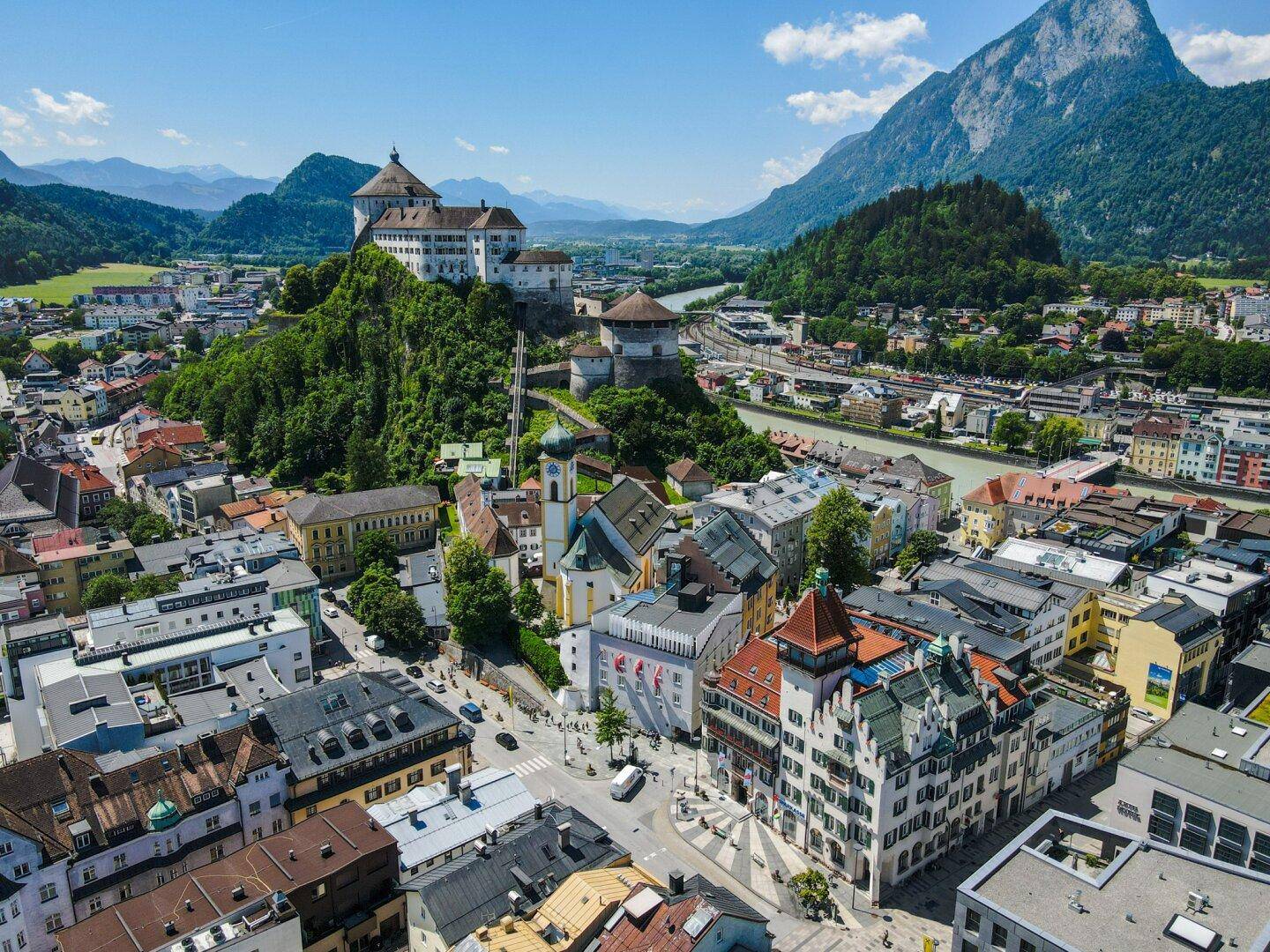 Panoramablick auf eine malerische Stadt mit historischer Burg, umgeben von majestätischen Bergen und grüner Landschaft.