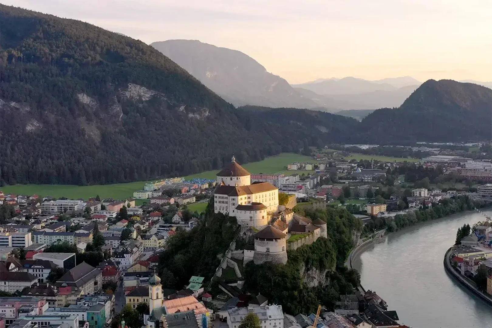 Luftaufnahme einer historischen Stadt mit einer imposanten Burg auf einem Hügel, umgeben von Bergen und einem Fluss.