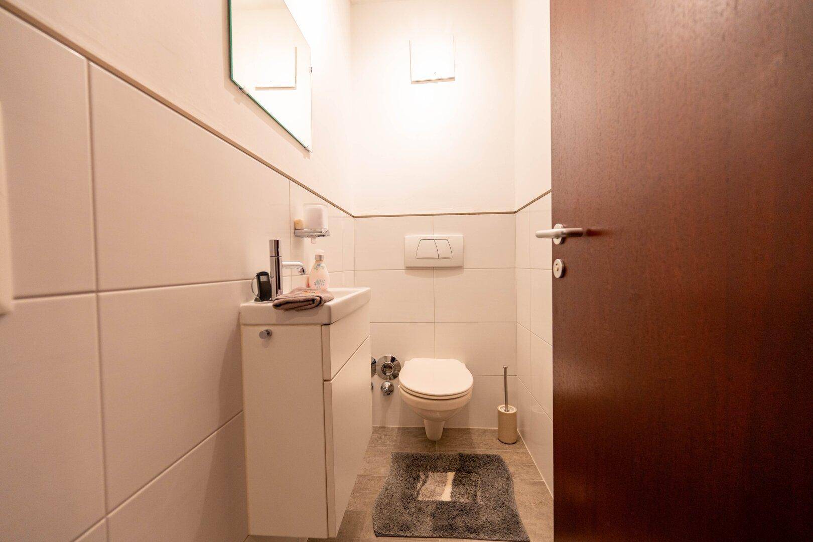 Kleines Gäste-WC mit Wand-WC, Handwaschbecken und hellen Fliesen.
