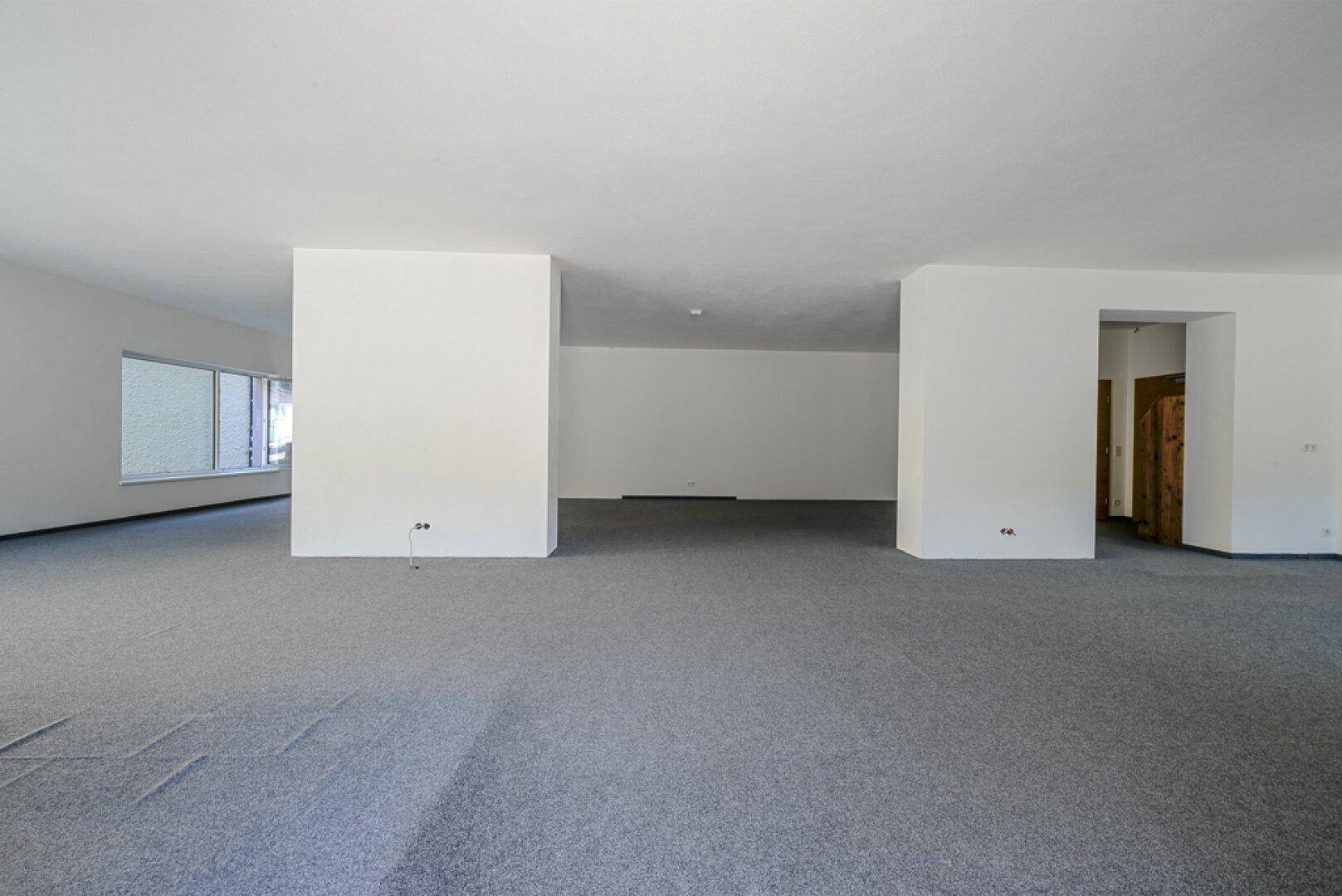 Großer, leerer Gewerberaum mit grauem Teppichboden und weißen Wänden, bereit zur Gestaltung.
