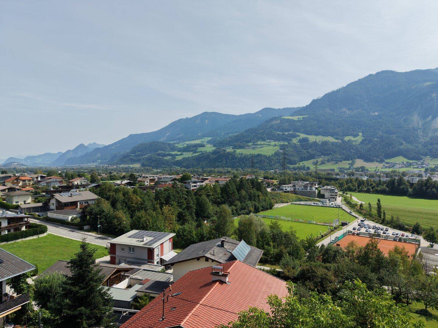 Weitläufige Luftaufnahme einer Stadt mit grünen Feldern und majestätischen Bergen im Hintergrund.