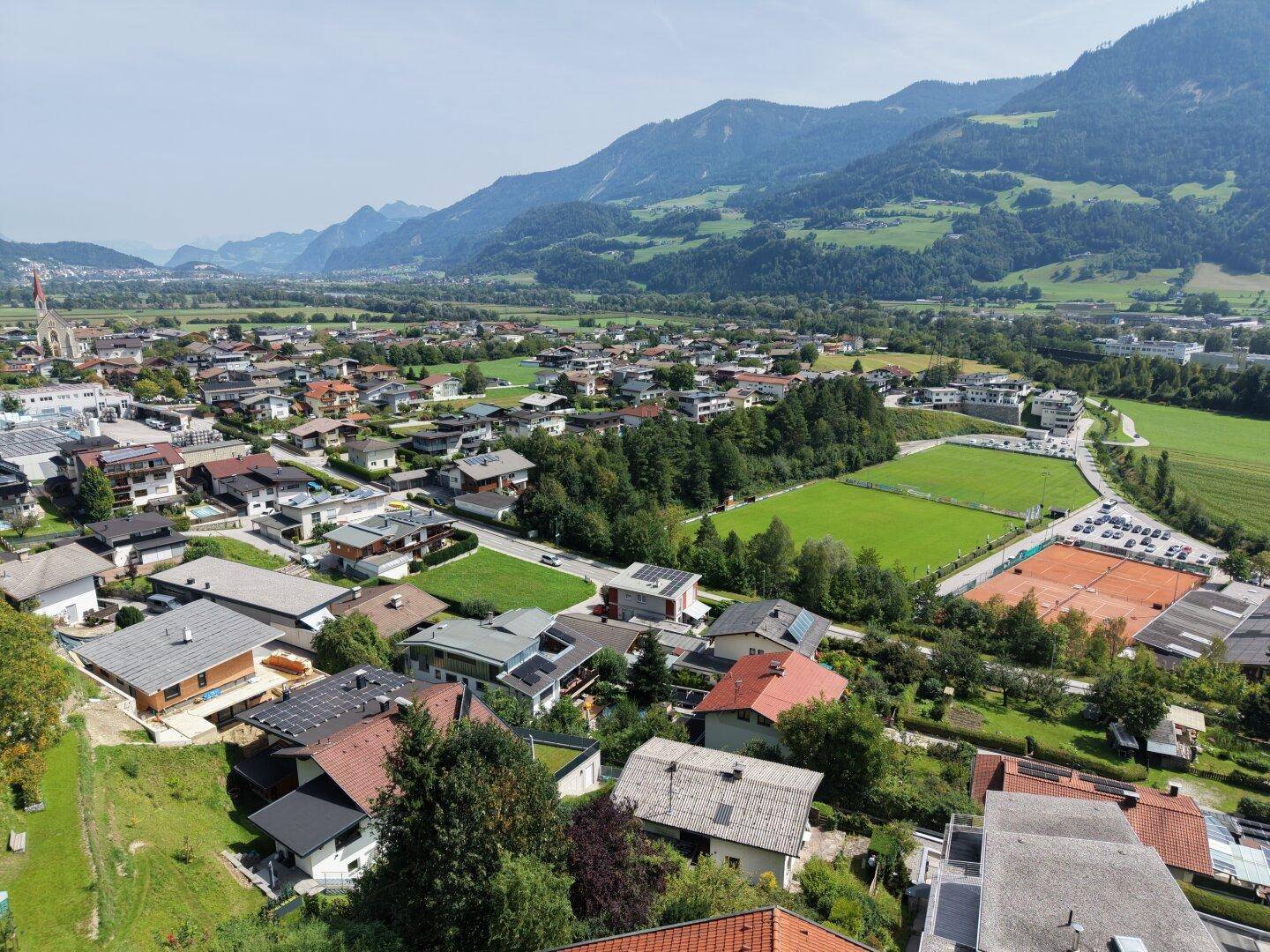 Weitwinkelaufnahme einer Siedlung mit grünen Wiesen, Sportanlagen und einer imposanten Bergkette.