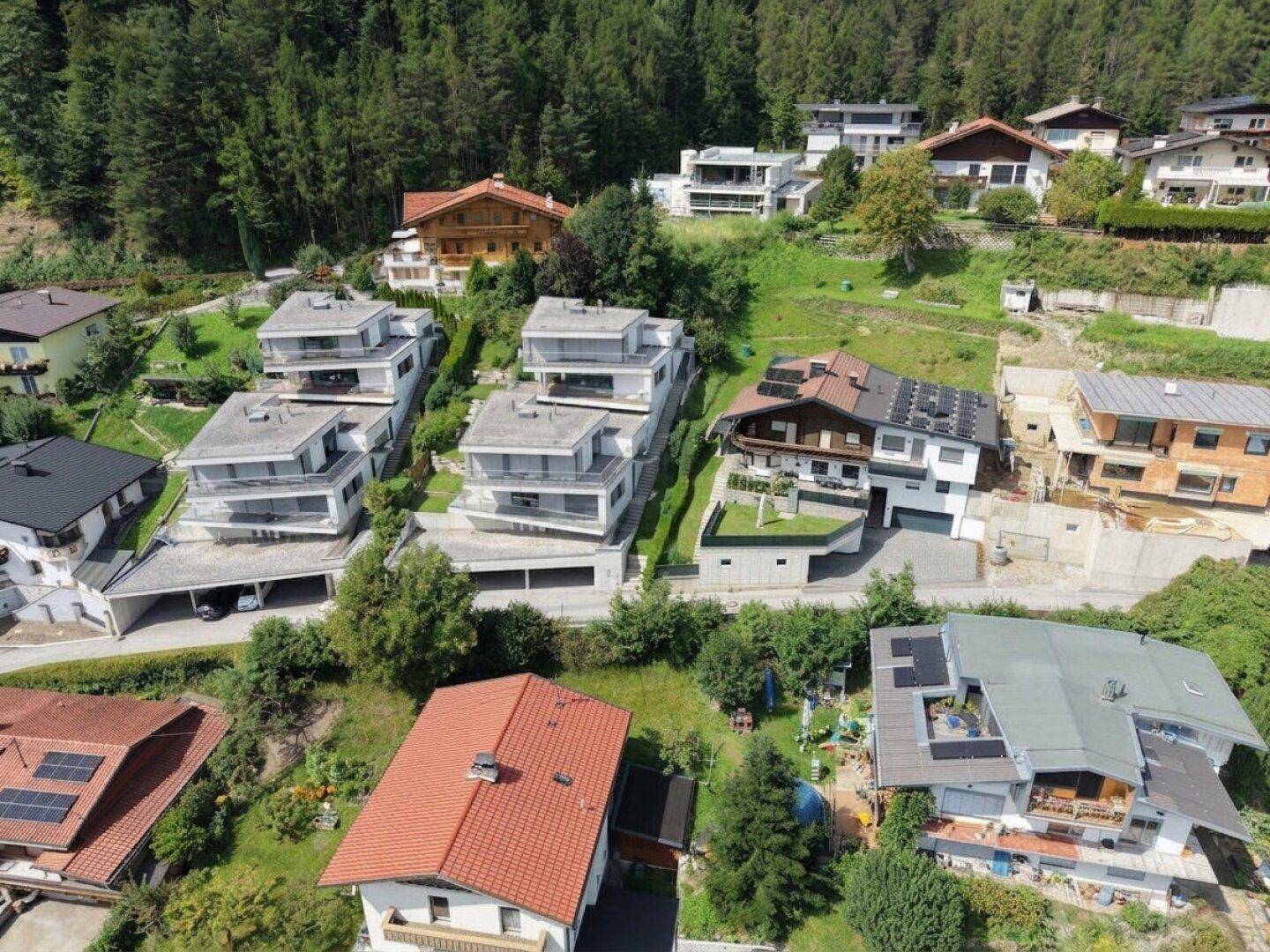 Luftaufnahme einer Wohnsiedlung am Hang mit modernen und traditionellen Häusern, umgeben von Wald.