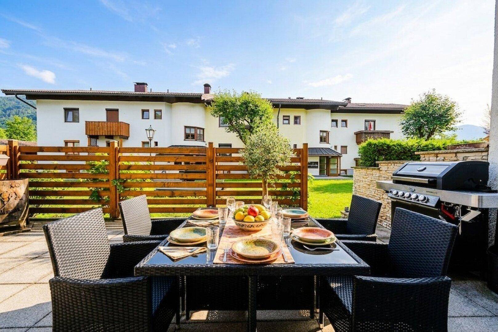Geräumige Terrasse mit dunklem Esstisch und Grill, umgeben von einem Holzzaun und Grünflächen.