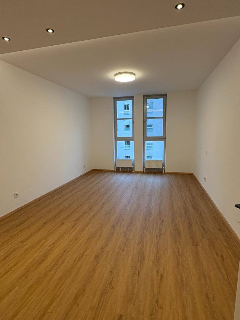 Heller, leerer Raum mit Laminatboden, weißen Wänden und hohen Fenstern für viel Tageslicht.