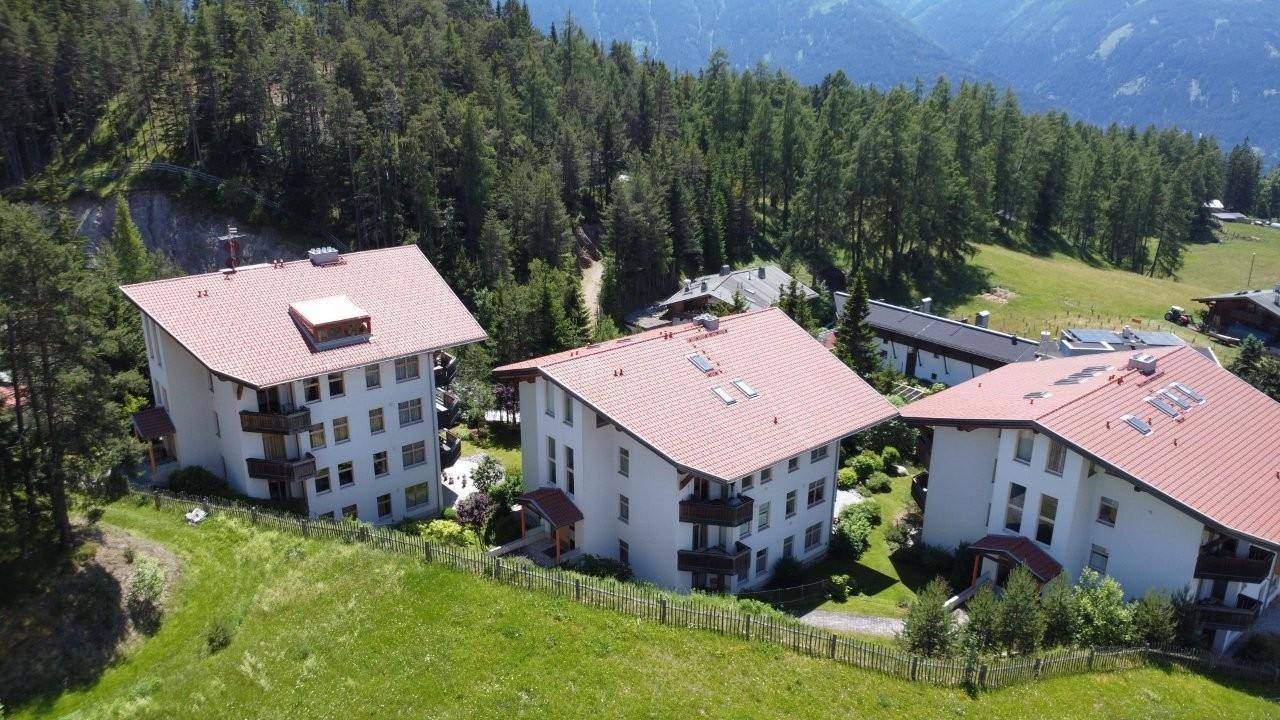 Drei ähnliche Gebäude mit roten Dächern und Balkonen, eingebettet in eine grüne Hügellandschaft mit Bergblick.
