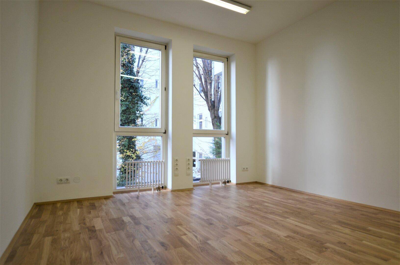 Heller Büroraum mit Holzboden und zwei hohen Fenstern, die viel Tageslicht hereinlassen.