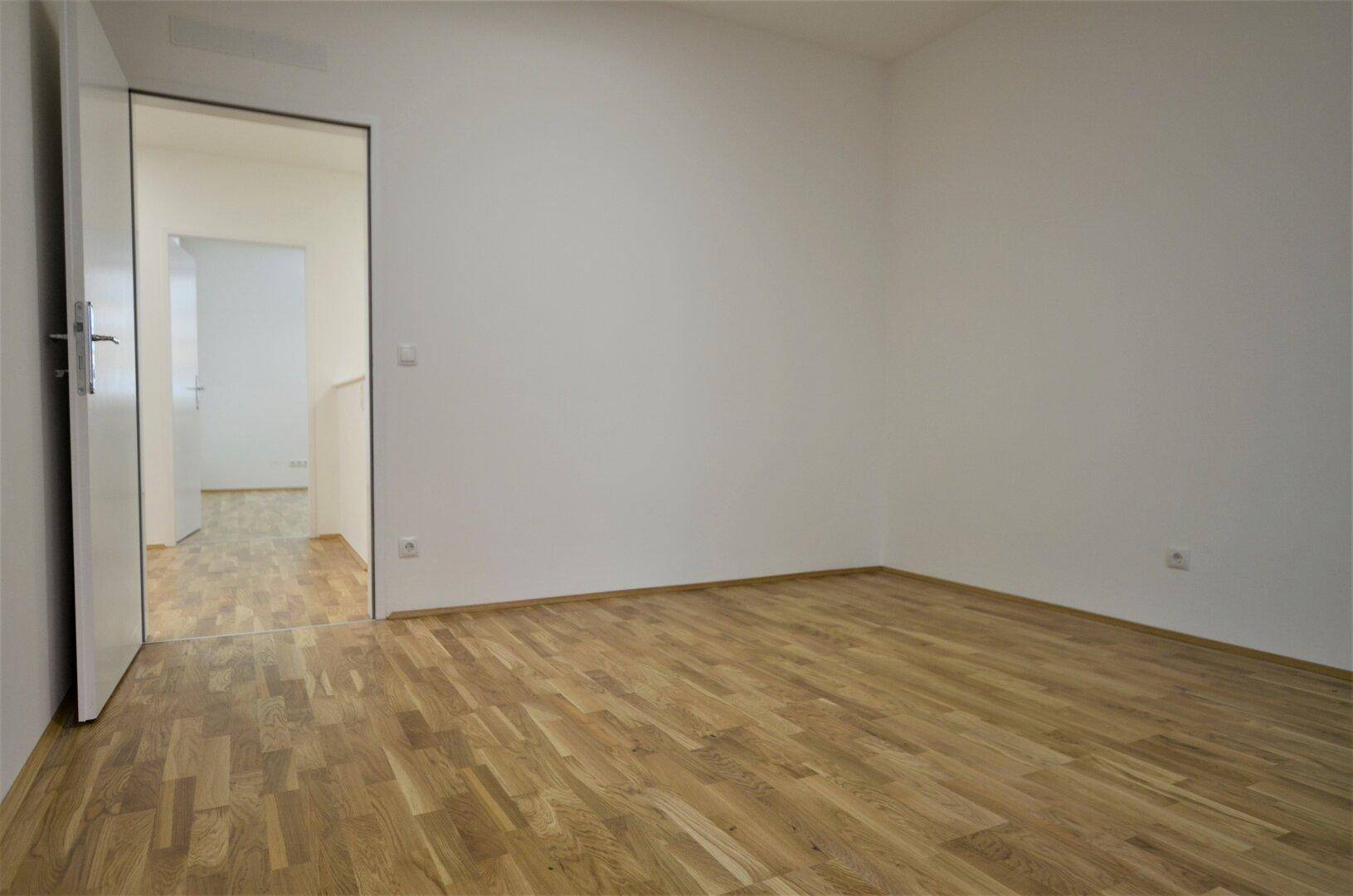 Schlichter Büroraum mit Holzboden und weiß gestrichenen Wänden, mit offener Tür zu einem weiteren Bereich.