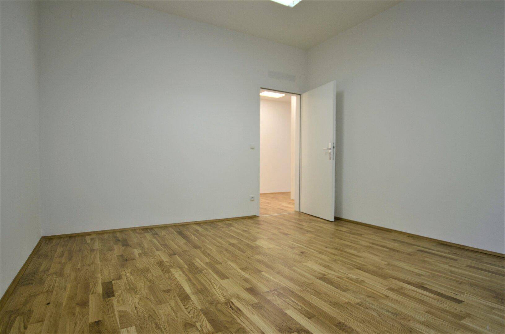 Leerer Büroraum mit Holzboden und weiß gestrichenen Wänden, mit offener Tür zu einem weiteren Bereich.