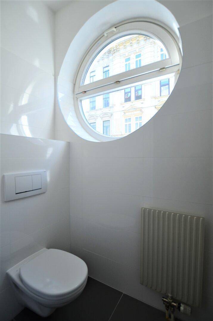 Modernes WC mit ovalem Fenster und Wand-WC, gefliest in Weiß und Grau.