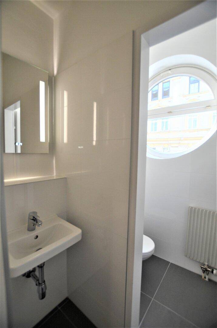 Modernes WC mit Waschbecken und Spiegel, durch eine Tür mit einem weiteren WC-Bereich verbunden.