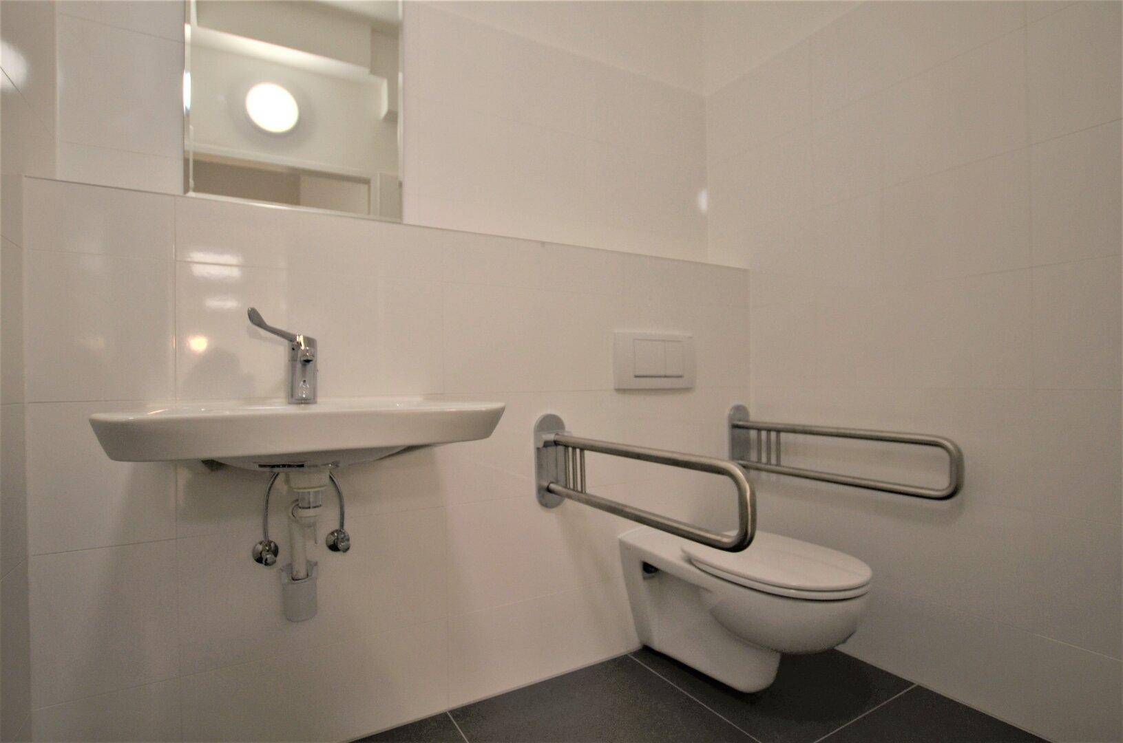Barrierefreies Badezimmer mit Haltegriffen am WC und modernem Waschbecken, gefliest in Weiß und Grau.