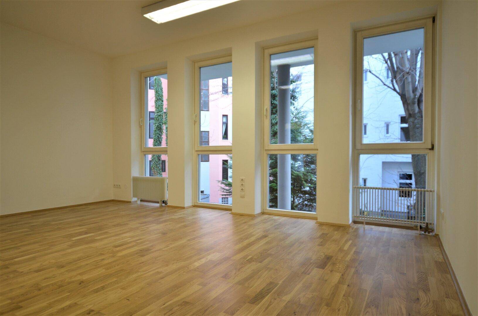 Heller Büroraum mit Holzboden und mehreren Fenstern, die einen Blick ins Grüne bieten.