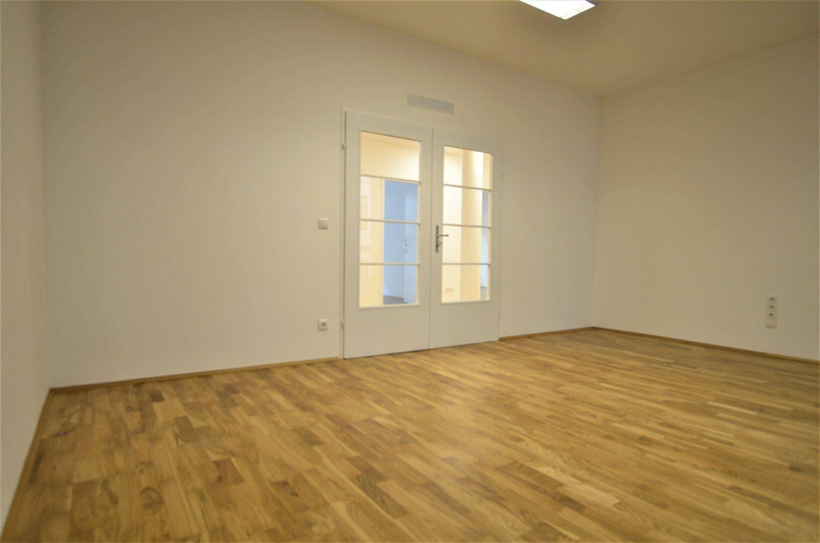 Heller Büroraum mit Holzboden und einer Doppeltür mit Glaseinsätzen, die zu einem weiteren Bereich führt.
