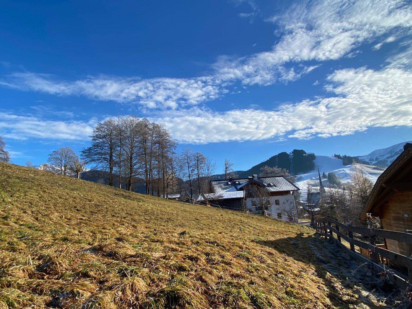 Sonniger Blick über eine Wiese auf ein Dorf mit traditionellen Häusern und schneebedeckten Bergen.