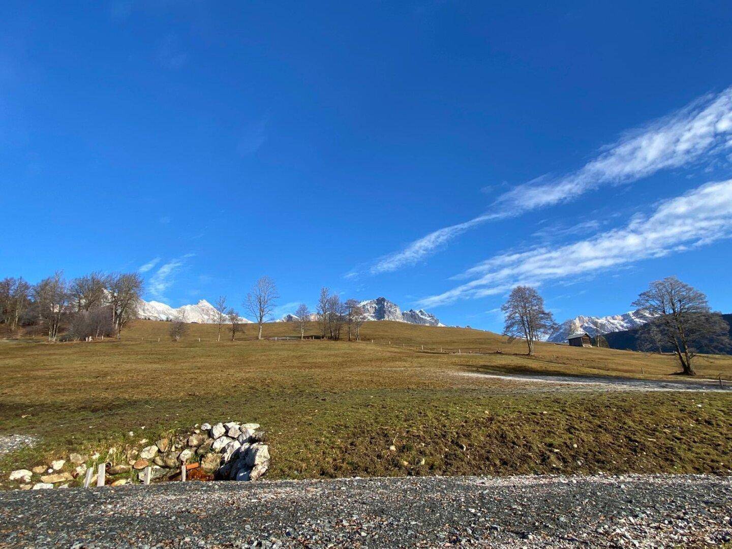 Panoramablick über eine ländliche Gegend mit Schotterweg, Wiesen und einer majestätischen Bergkulisse.