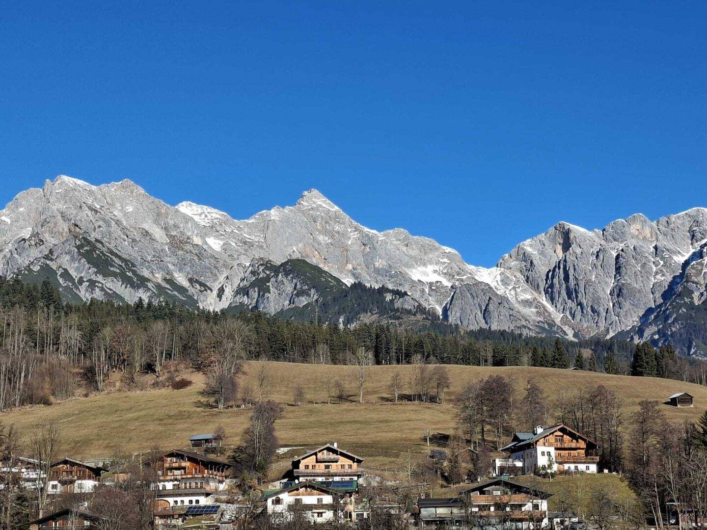 Weitläufiger Blick auf ein Tal mit Häusern am Hang und imposanten schneebedeckten Bergen im Hintergrund.