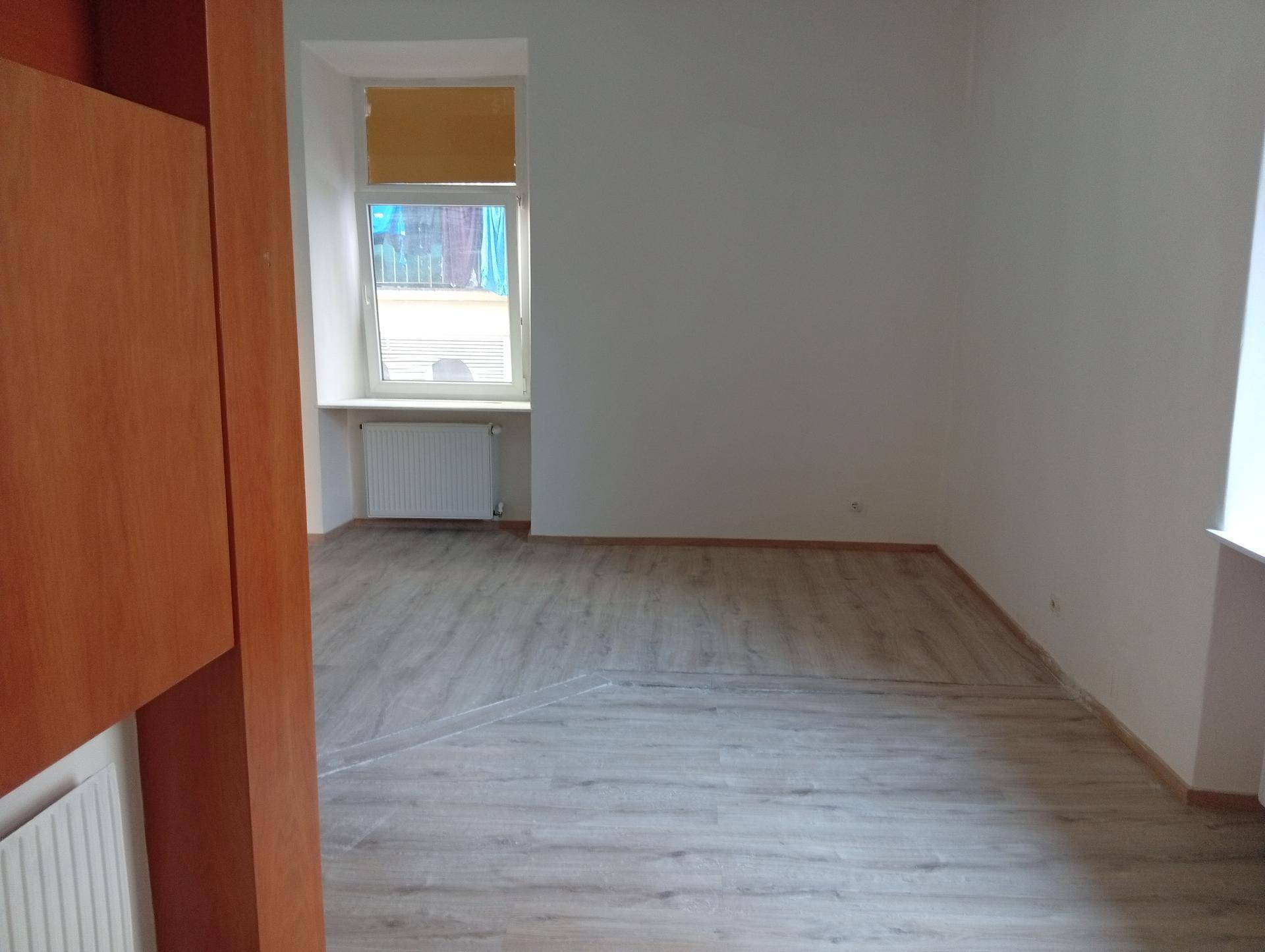 Leerer Raum mit hellem Laminatboden, weiß gestrichenen Wänden und einem Fenster mit Blick nach draußen.
