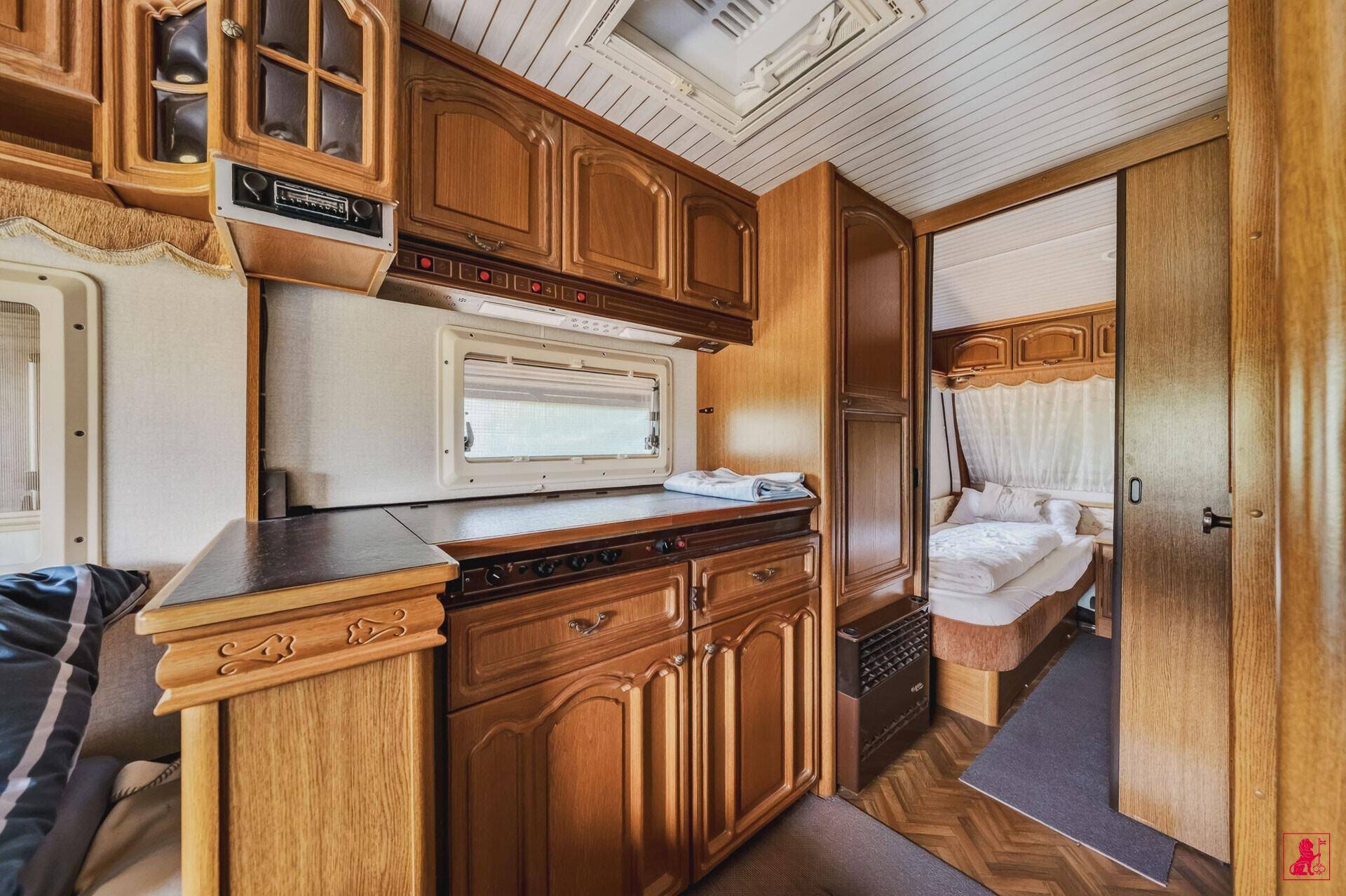 Kleine Küchenzeile in einem Wohnwagen mit Holzschränken und Blick in ein weiteres Schlafzimmer.