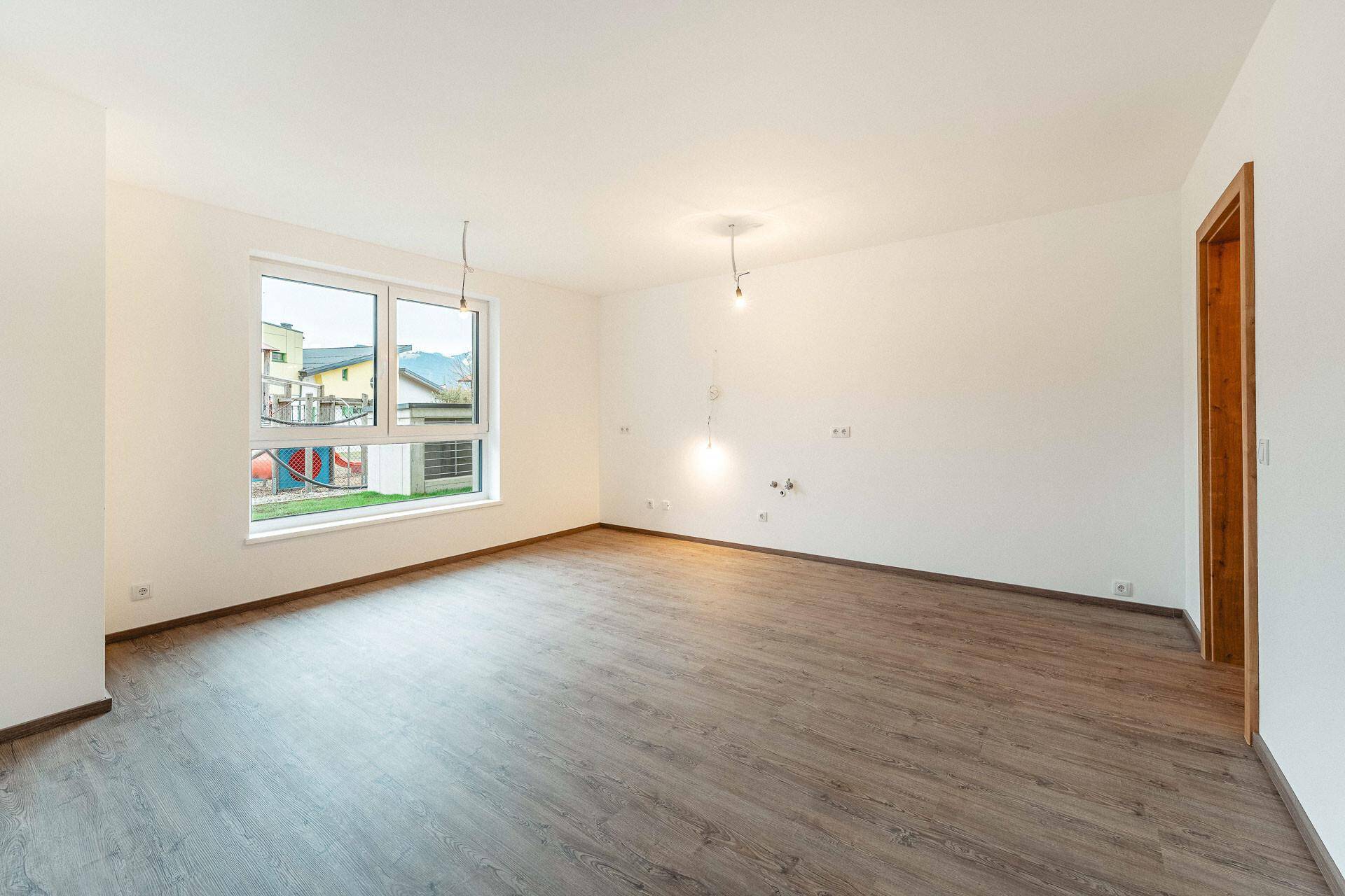 Helles, leeres Zimmer mit modernem Laminatboden und Blick auf einen Spielplatz.