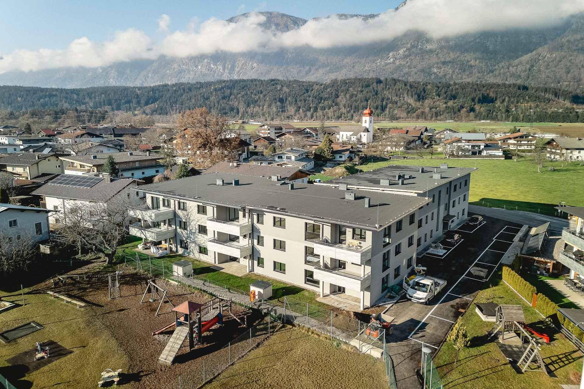 Luftaufnahme eines modernen Mehrfamilienhauses mit Spielplatz und Bergblick in ländlicher Umgebung.