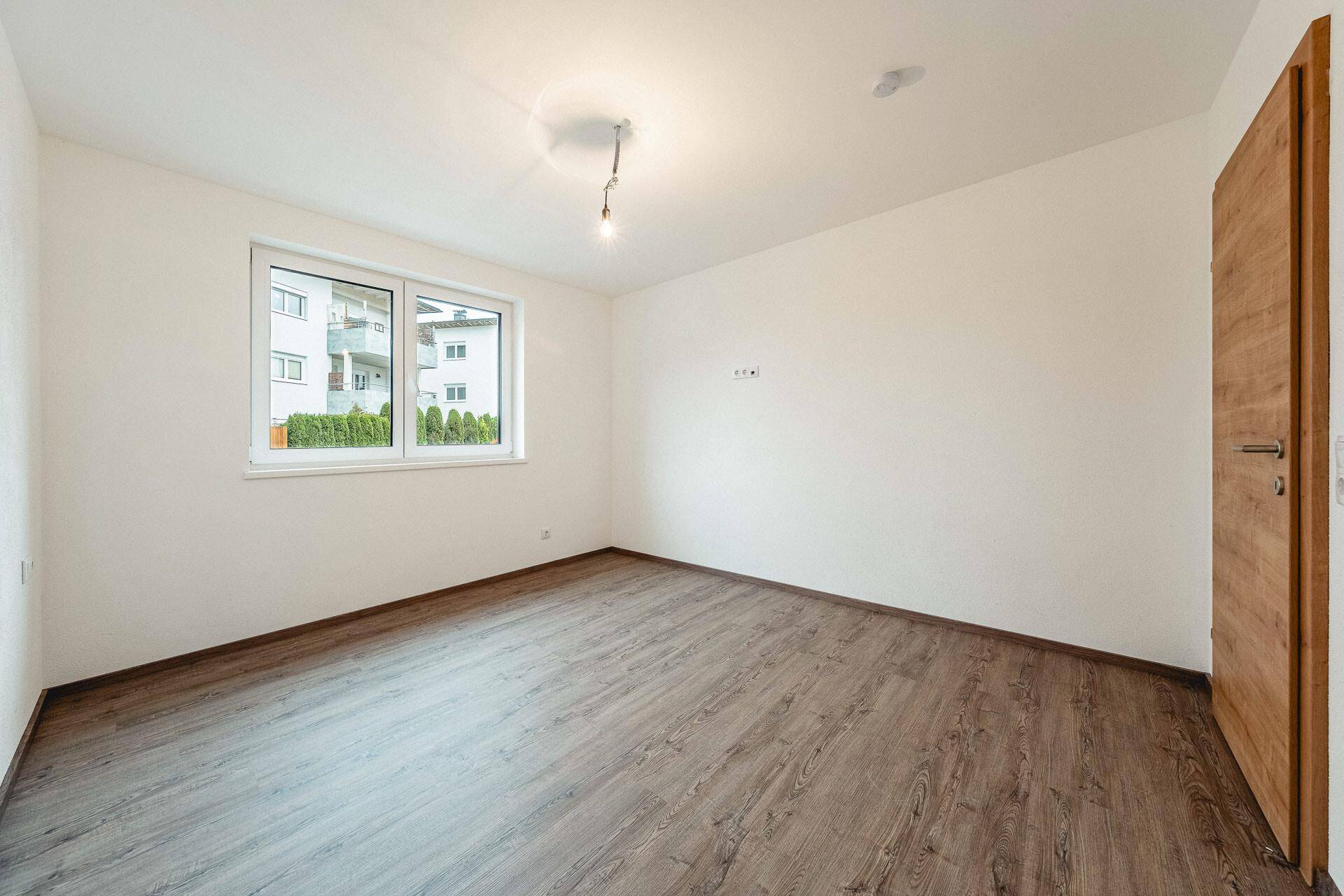 Unmöbliertes Schlafzimmer mit hellem Laminatboden und Fenster, das viel Tageslicht hereinlässt.