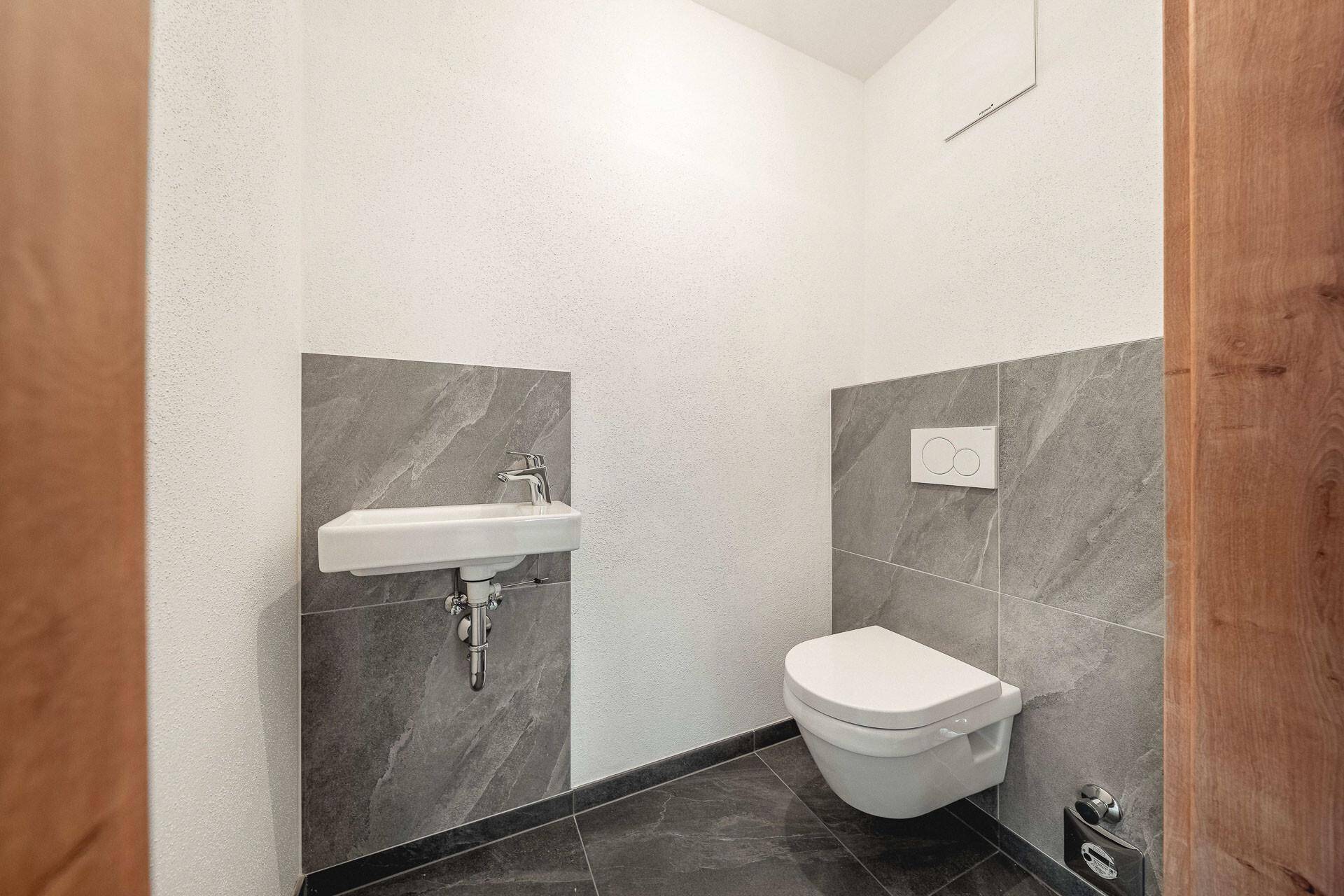 Modernes Gäste-WC mit wandhängender Toilette, kleinem Waschbecken und grauen Fliesen.