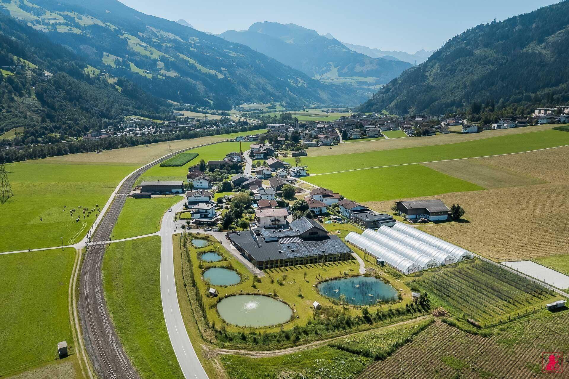 Panoramablick auf die weitläufige Anlage mit Teichen und Gewächshäusern, umgeben von einem Dorf und Bergen.