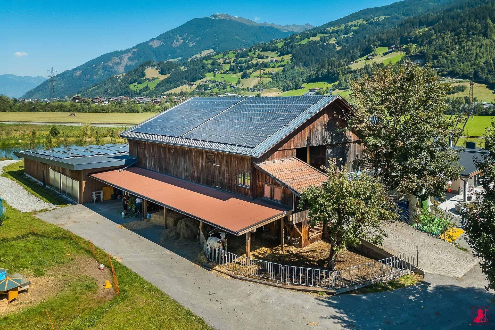 Rustikales Scheunengebäude mit Photovoltaikanlage auf dem Dach, umgeben von grünen Wiesen und Bergen.