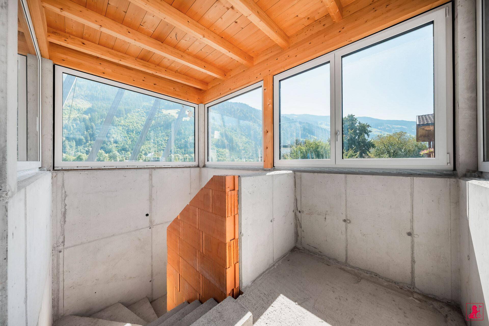 Unfertiger Raum mit großen Fenstern, die einen weiten Blick auf die Berge bieten, und einer Holzbalkendecke.