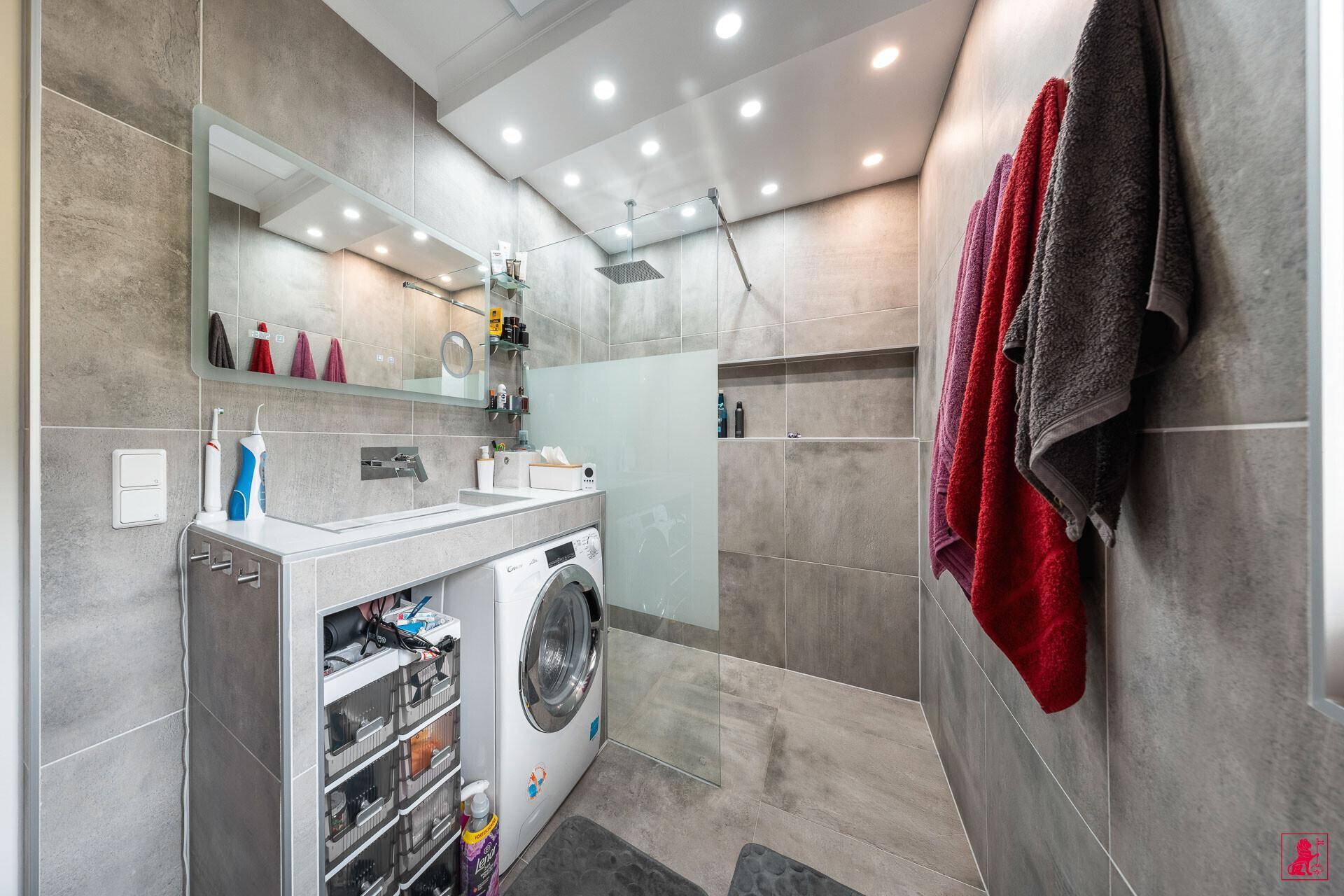 Modernes Badezimmer mit großer Dusche, Waschmaschine und grauen Fliesen.