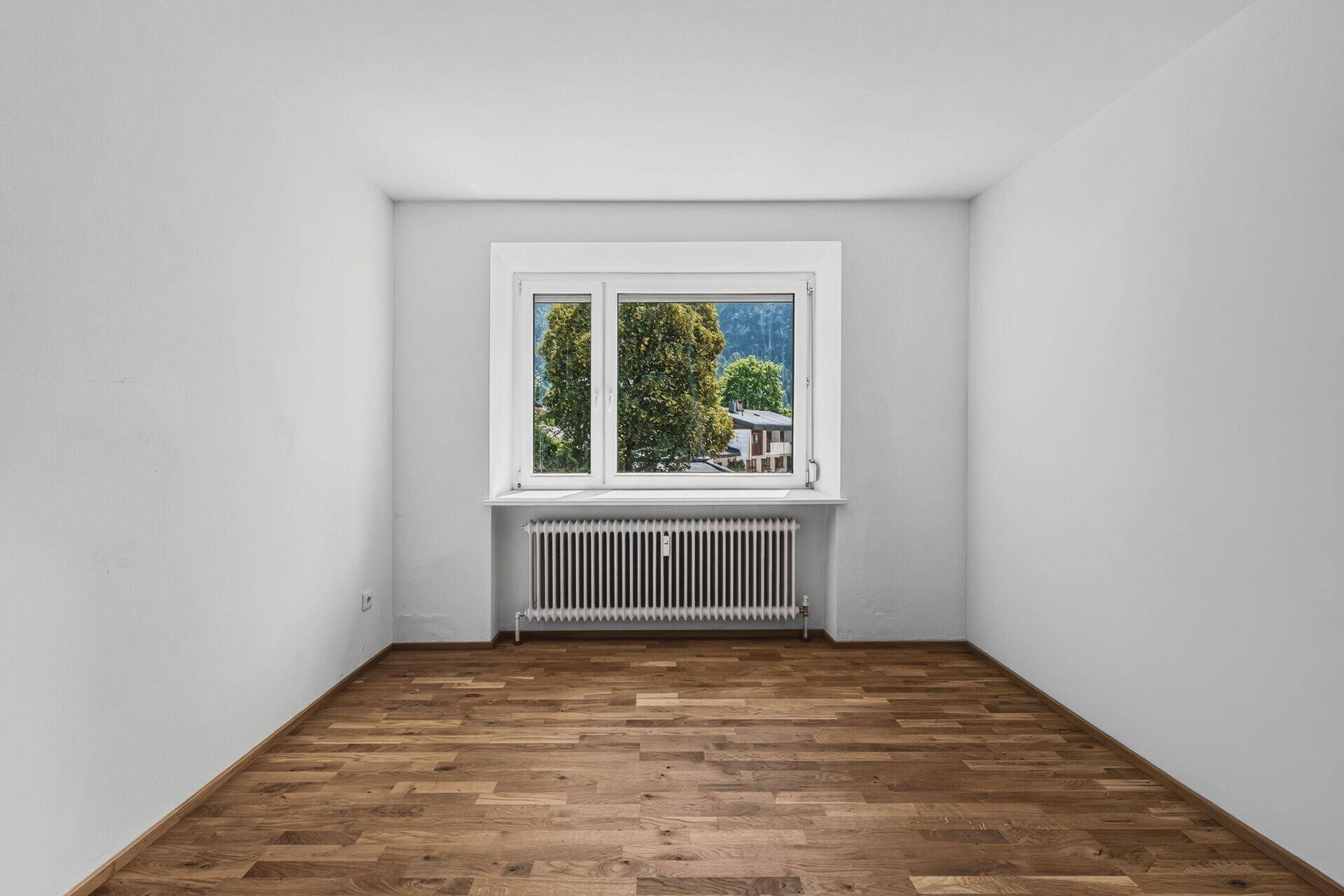 Kleines, helles Zimmer mit Parkettboden und einem Fenster mit Blick auf Bäume und Häuser.