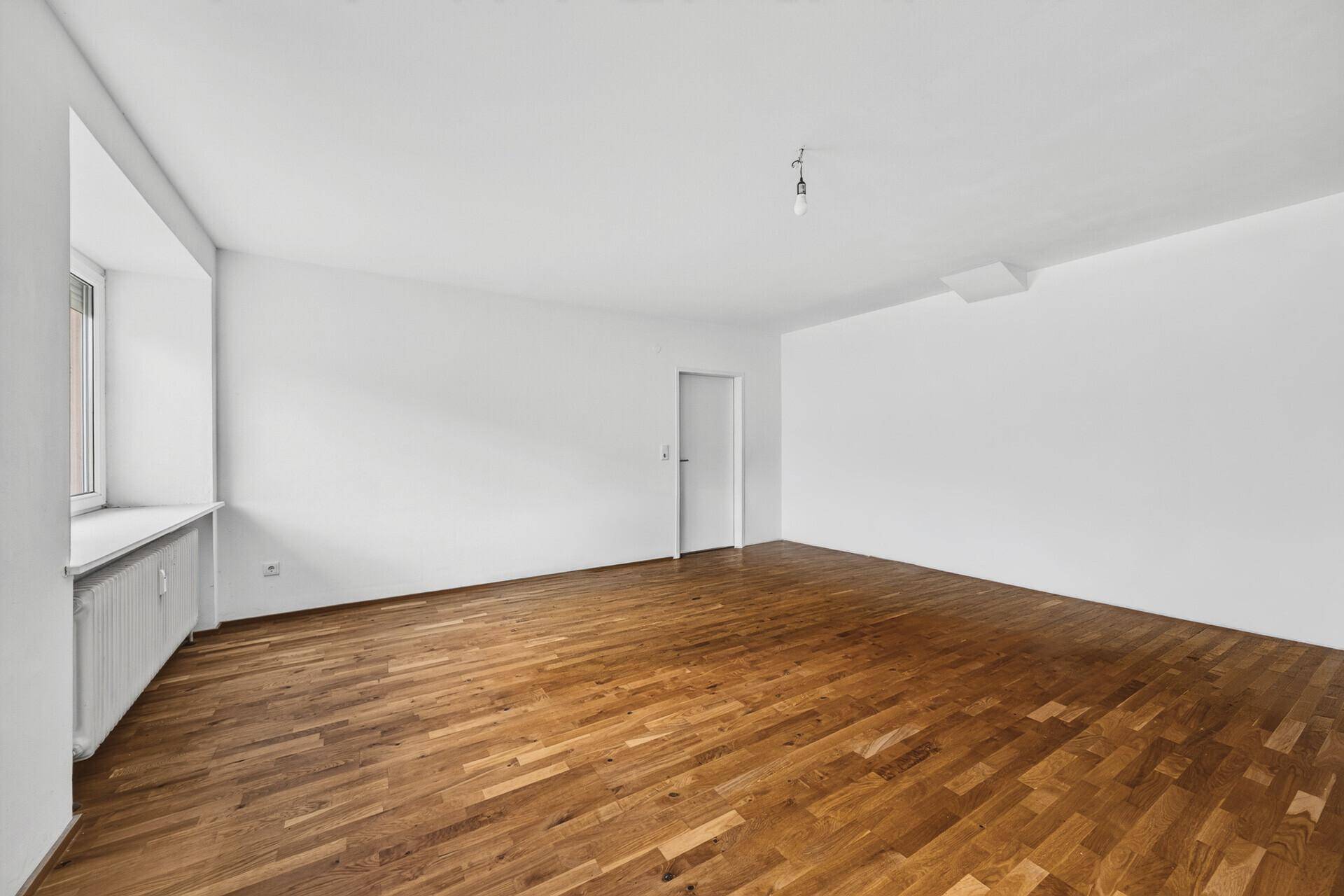 Geräumiges, leeres Zimmer mit Holzboden und einem Fenster, das viel Tageslicht hereinlässt.