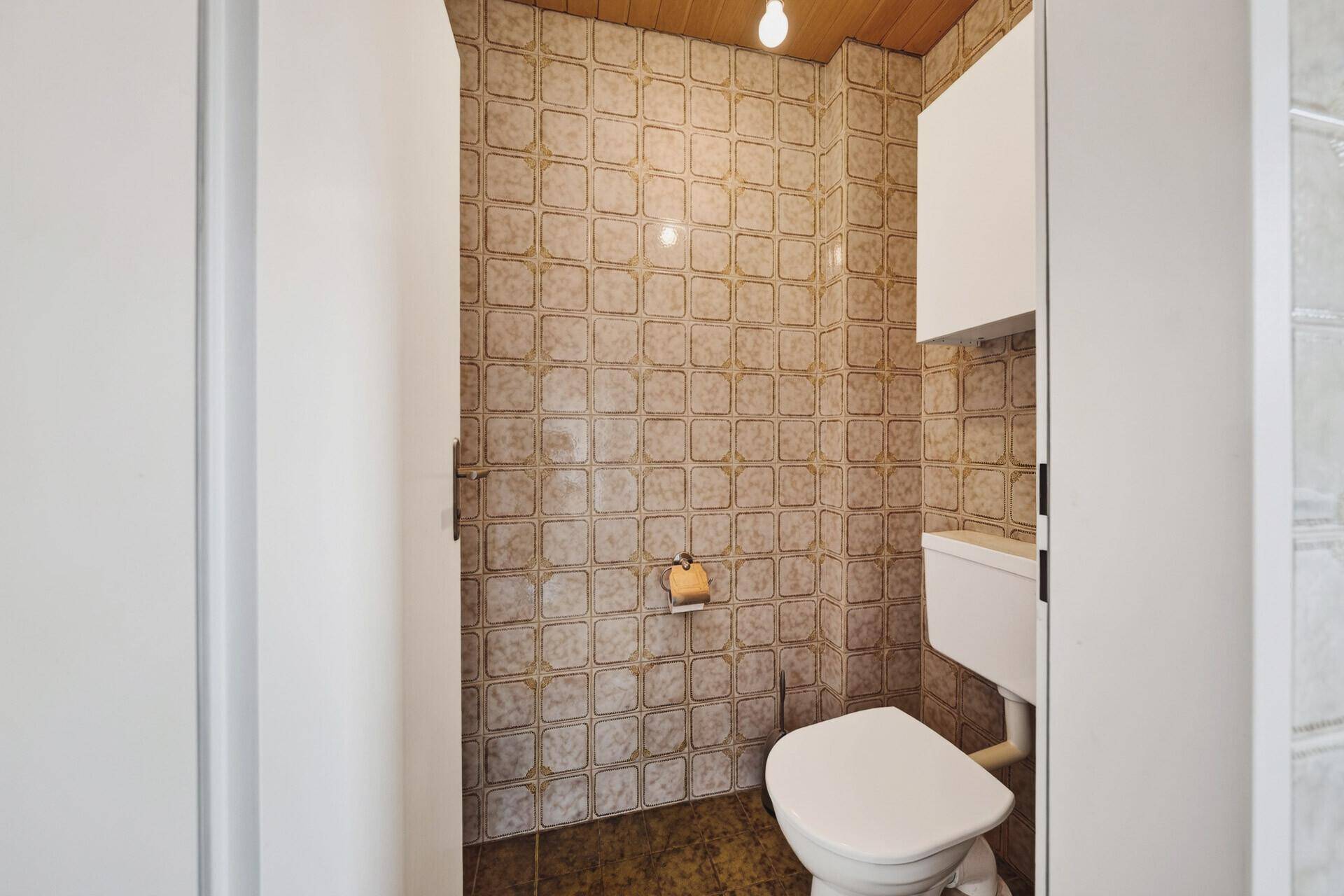 Separate Toilette mit Fliesen an Wänden und Boden sowie einer Holzdecke.