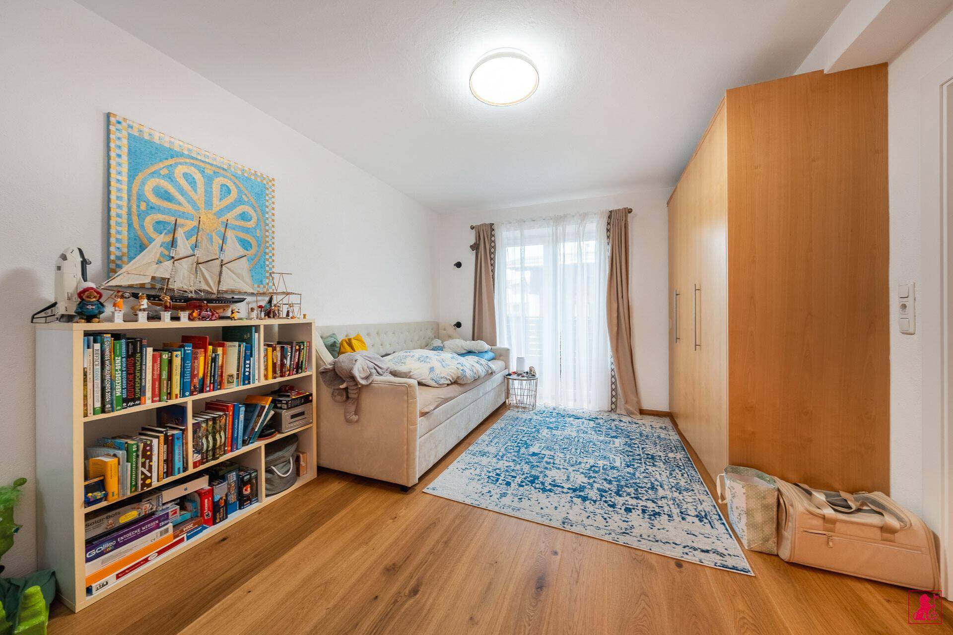 Helles Kinder- oder Gästezimmer mit Holzboden, Bücherregal und Zugang zum Balkon.