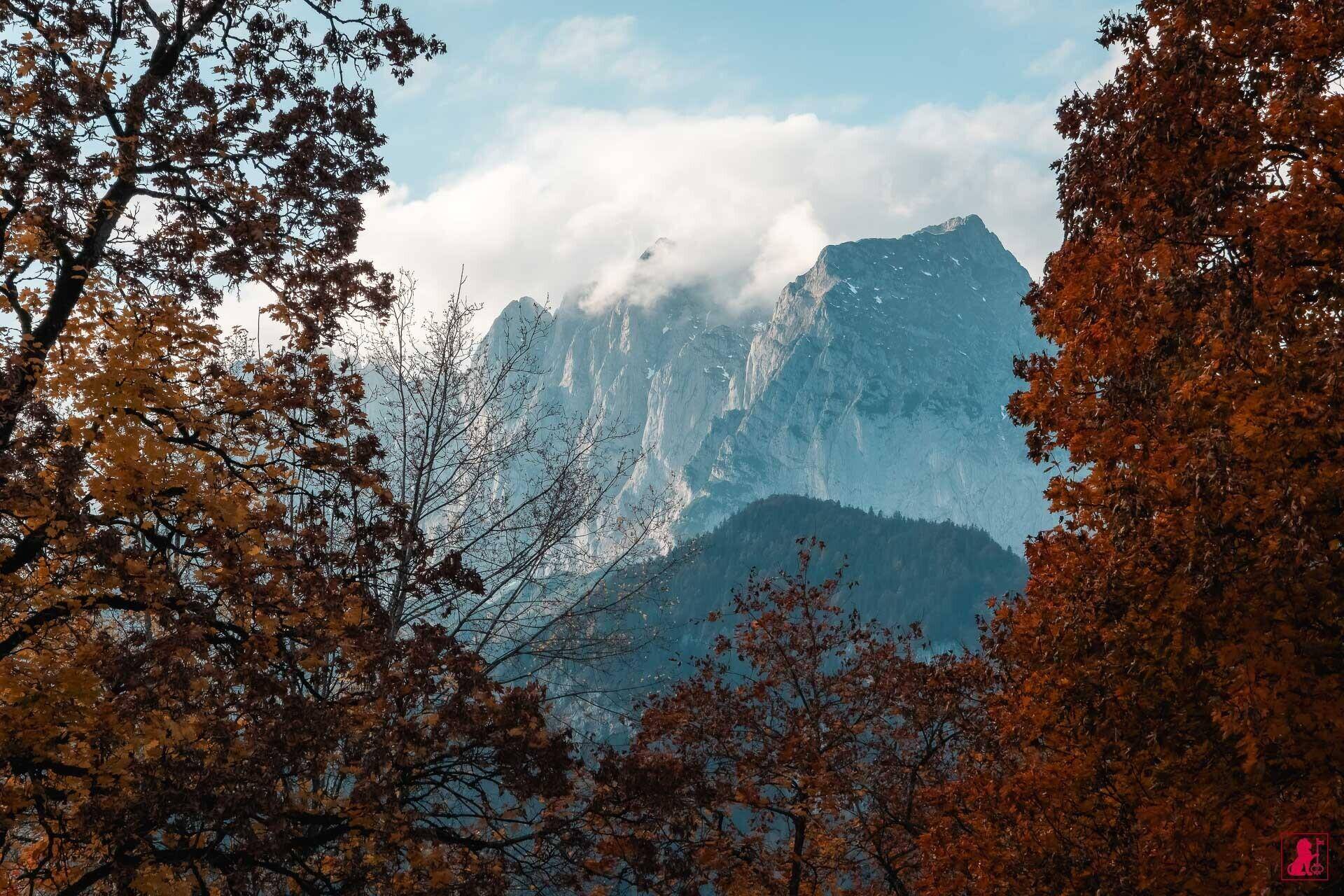 Malerischer Blick auf majestätische Berge, umrahmt von bunten Herbstbäumen unter einem bewölkten Himmel.