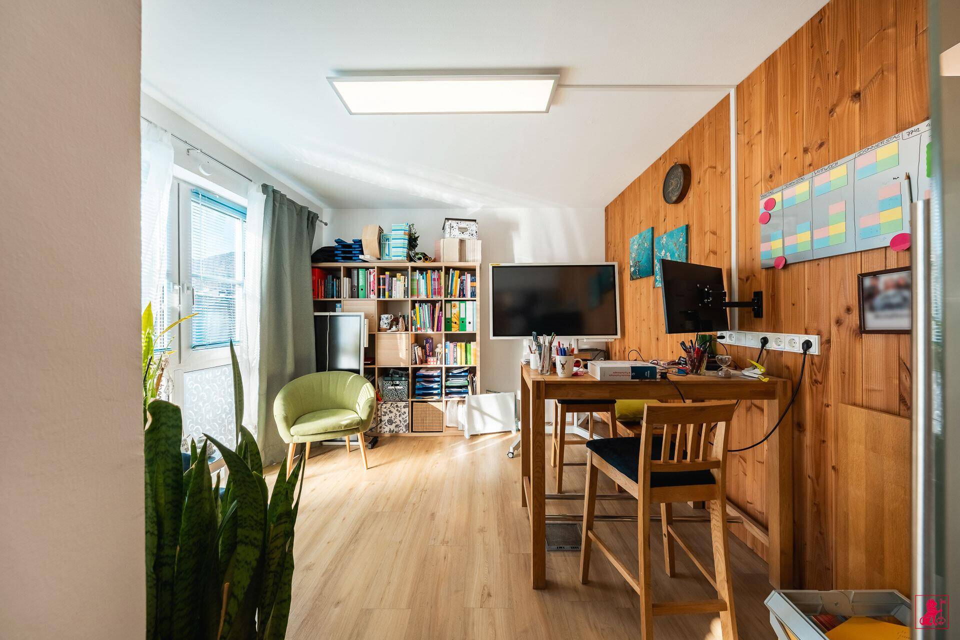 Heller Arbeitsplatz mit Schreibtisch, Bürostuhl und Bücherregal, gut beleuchtet durch große Fenster.