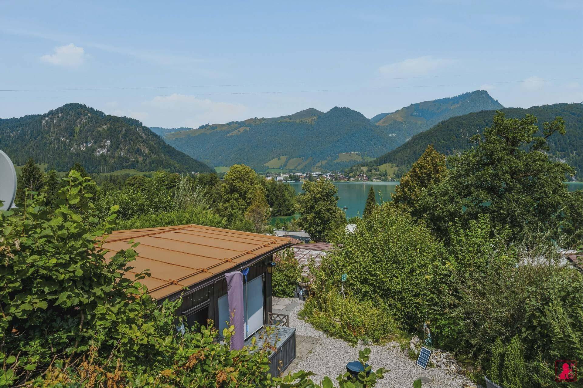 Blick über die Dächer und grüne Landschaft zum See und den umliegenden Bergen.