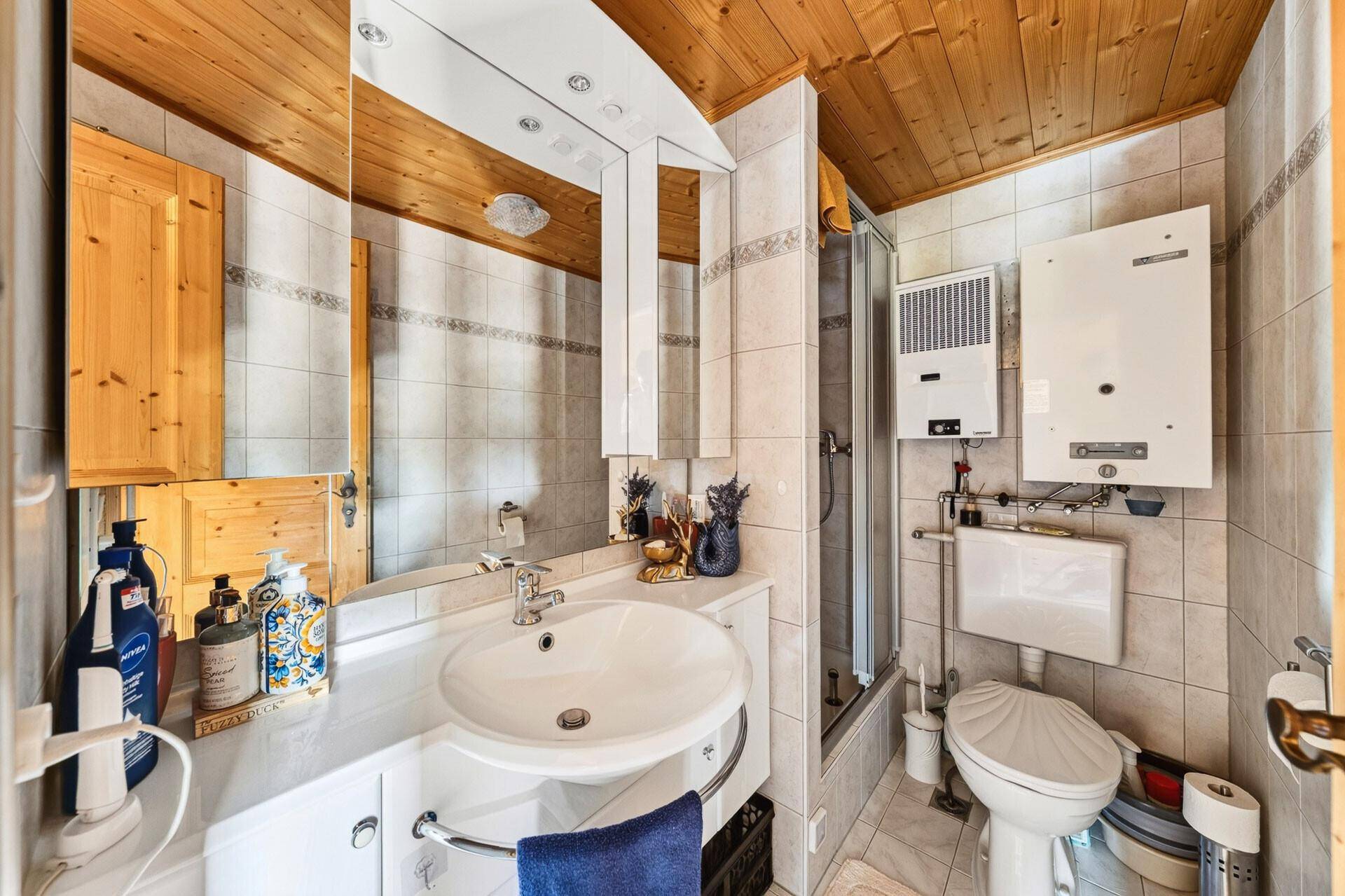 Kleines Badezimmer mit Dusche, Waschbecken und WC, komplett gefliest und mit Holzdecke.
