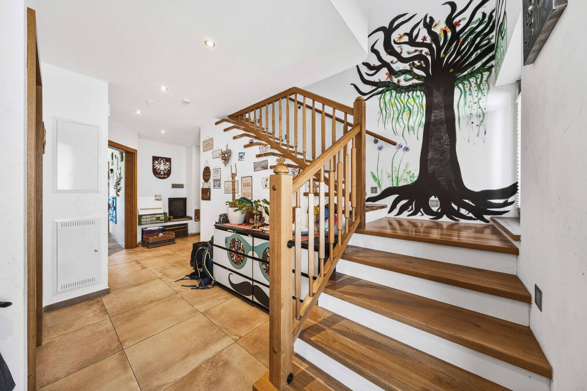 Heller Flur mit Holztreppe und einer auffälligen Baum-Wandmalerei, die zum Obergeschoss führt.