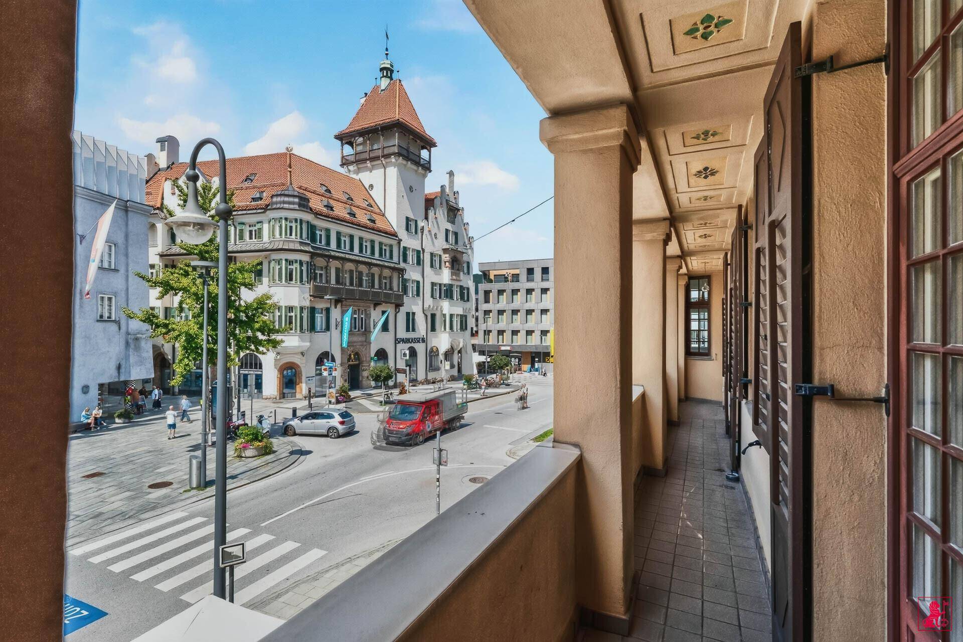 Blick von einer Loggia auf einen belebten Stadtplatz mit historischen Gebäuden und Passanten.