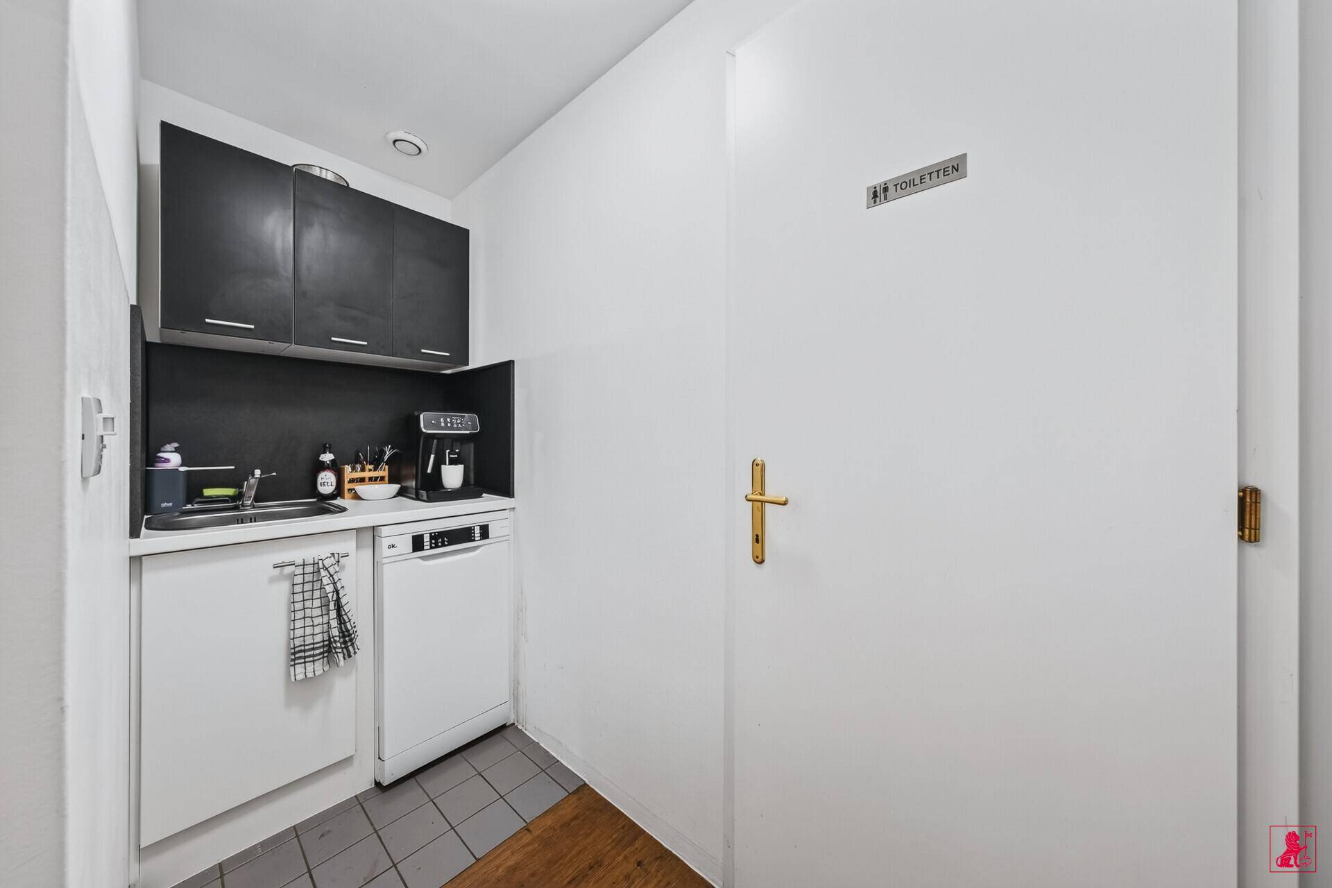 Kleine Teeküche mit Spülmaschine und Kaffeemaschine, angrenzend an einen Toilettenbereich.